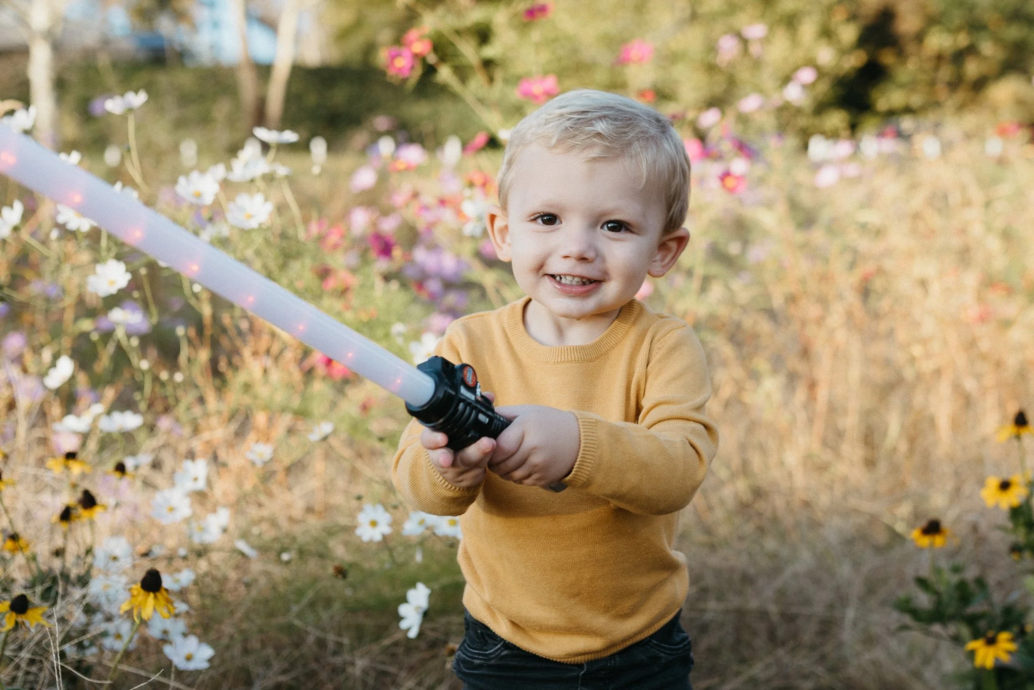 A young boy with blonde hair, wearing a yellow long-sleeved shirt and dark pants, holding a toy sword, standing outdoors among colorful flowers and trees, smiling at the camera.