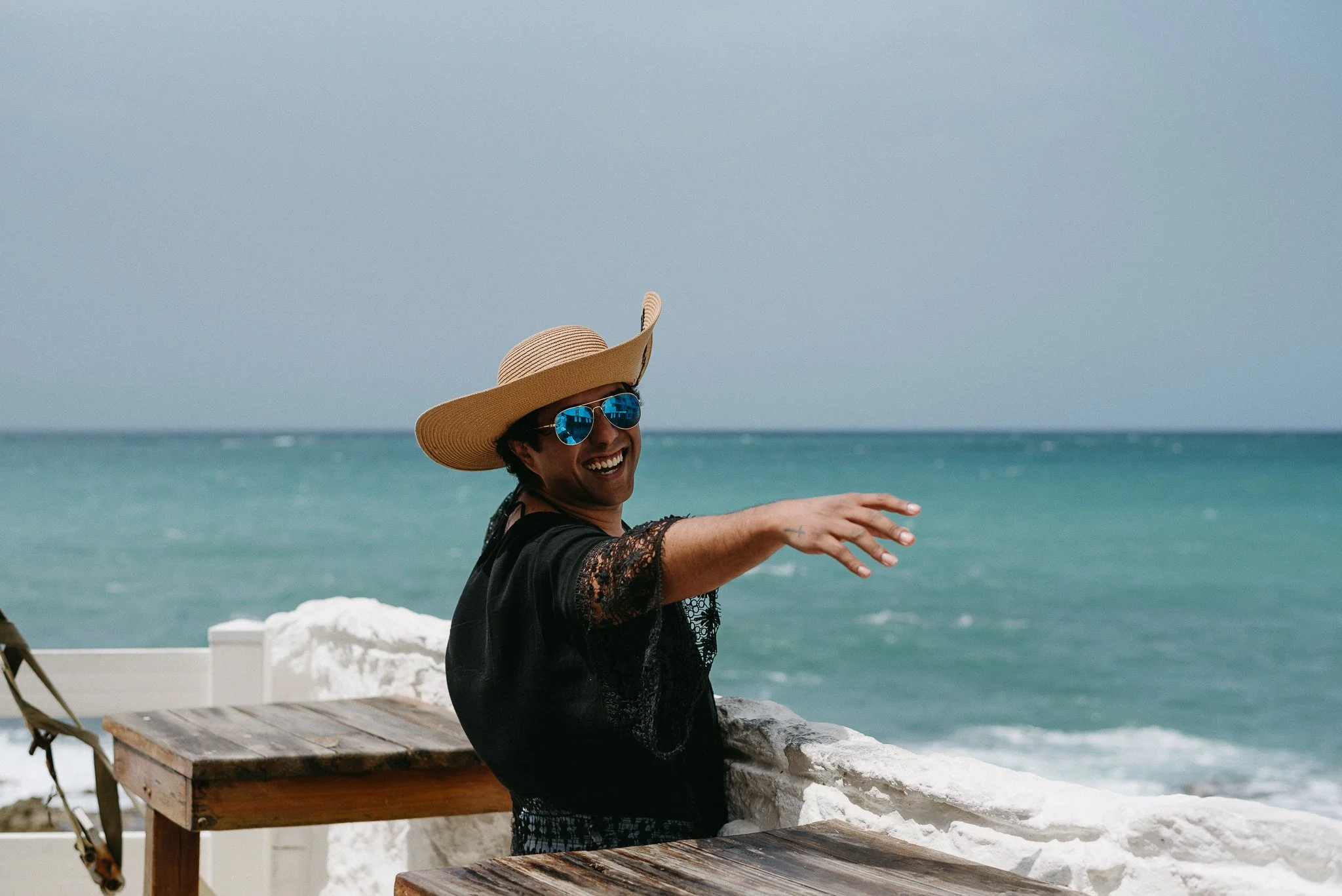 Person wearing a large straw hat and sunglasses with blue reflective lenses, smiling and pointing at the camera, sitting by the ocean.