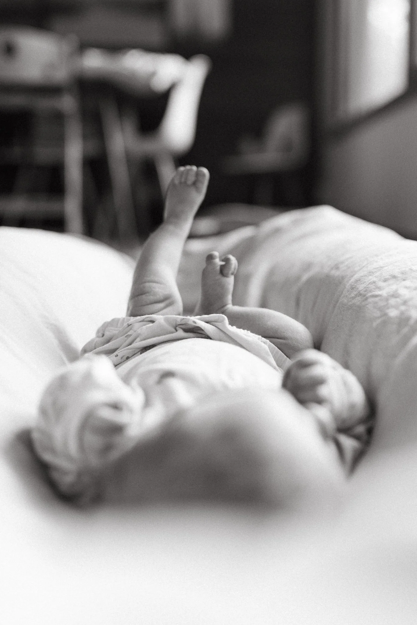 Black and white photograph of a newborn baby lying on a soft surface, with a person's hand reaching out in the background.