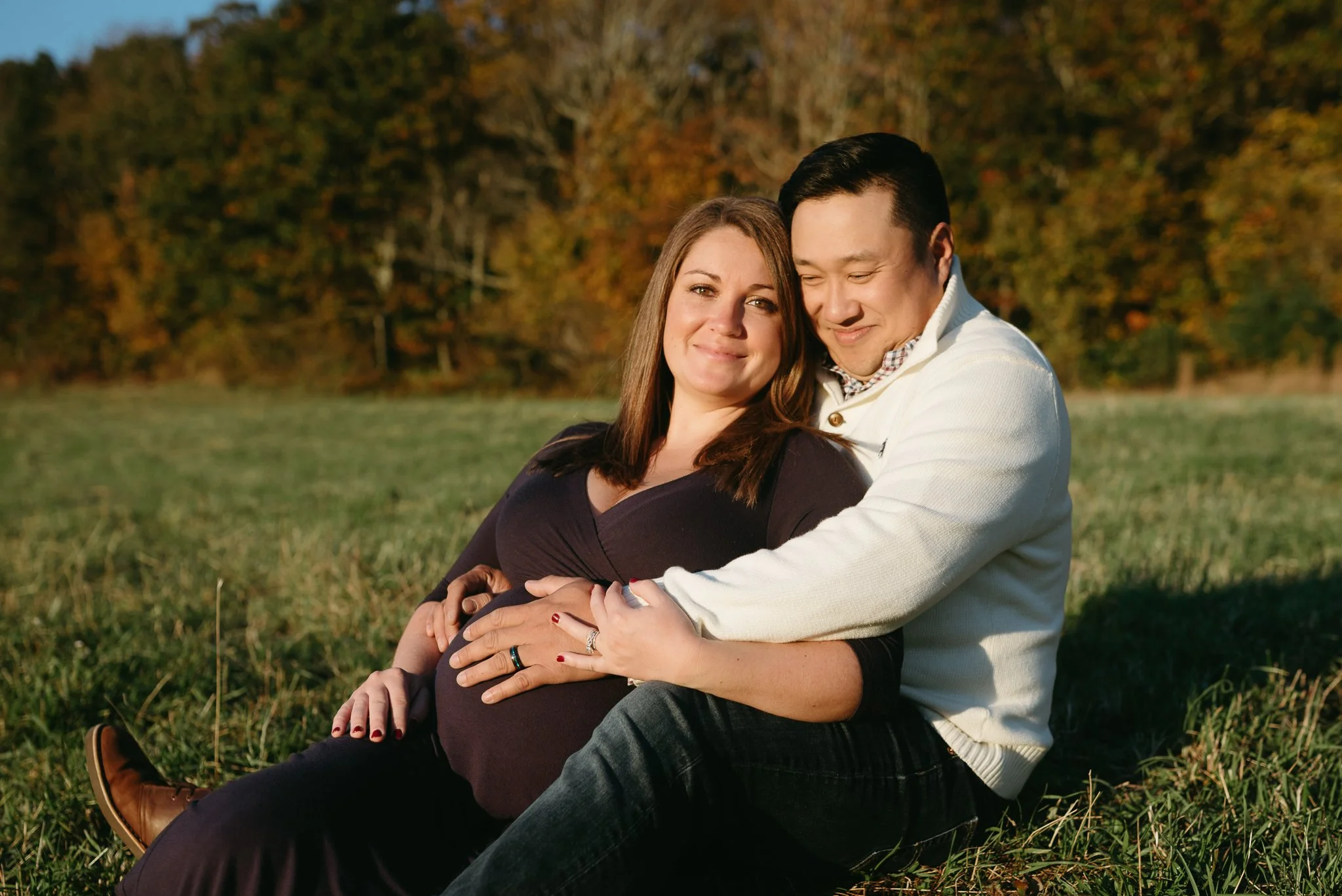 A couple sitting on the grass in a park with trees in the background, the woman showing a pregnant belly and the man embracing her, during sunset.
