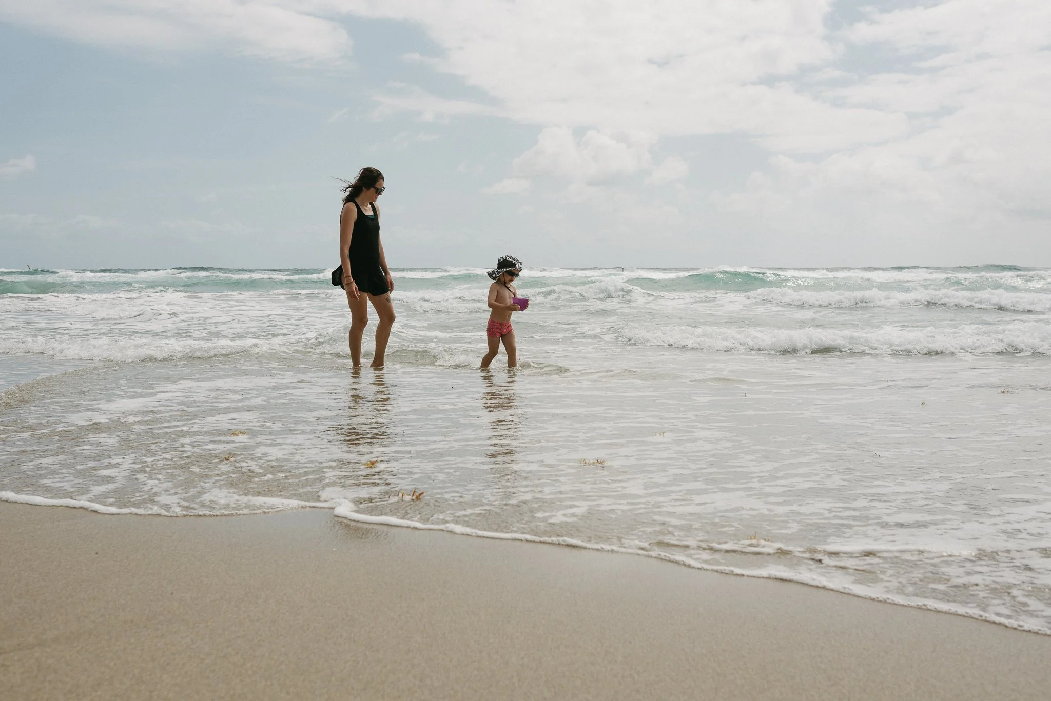 A woman and a young girl walking along the shoreline at the beach, with waves in the background and a partly cloudy sky.