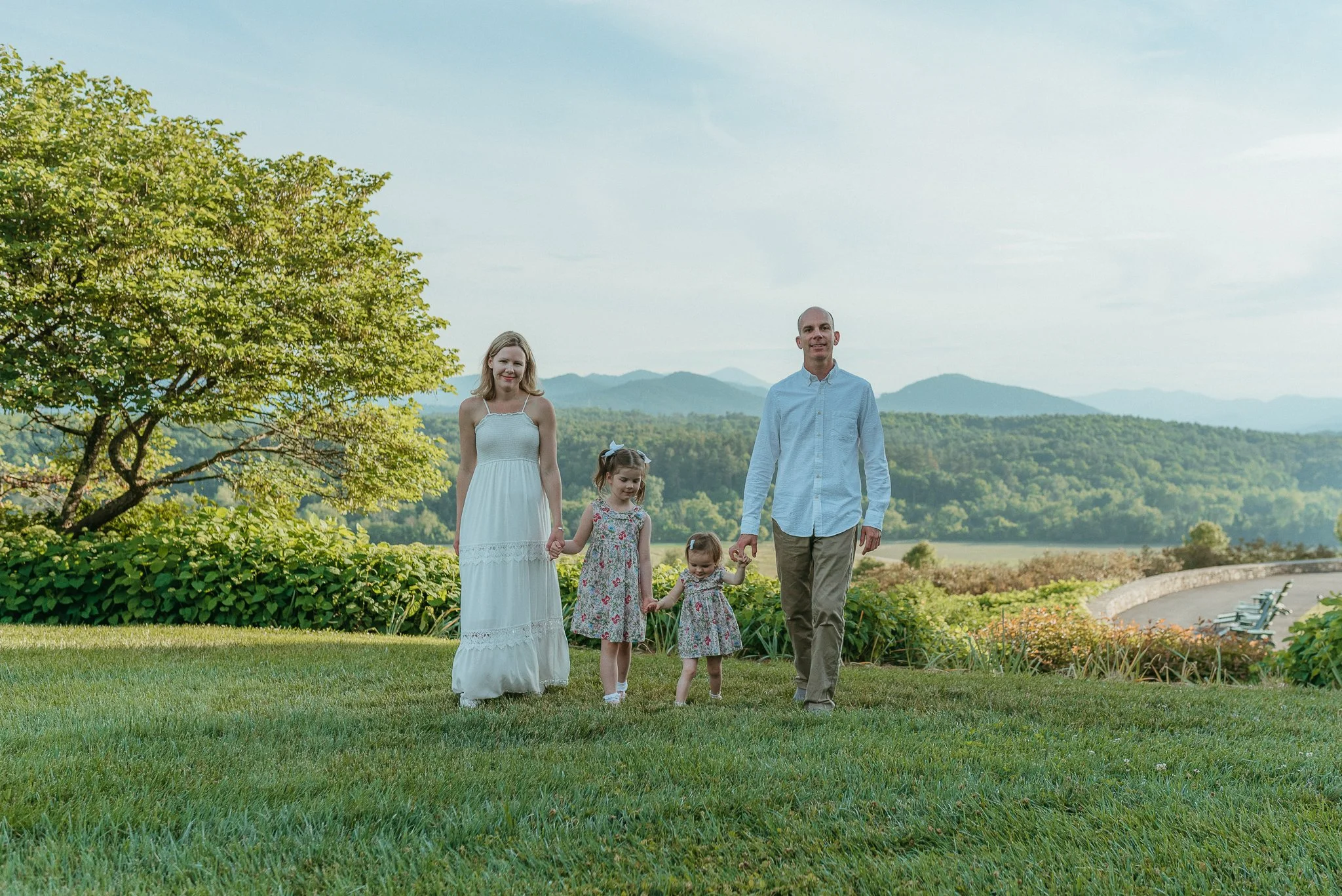 A family of six walking outdoors on a grassy area with mountains in the background. The family includes two women, one man, and three young girls. They are all dressed in light, summery clothing and appear happy.