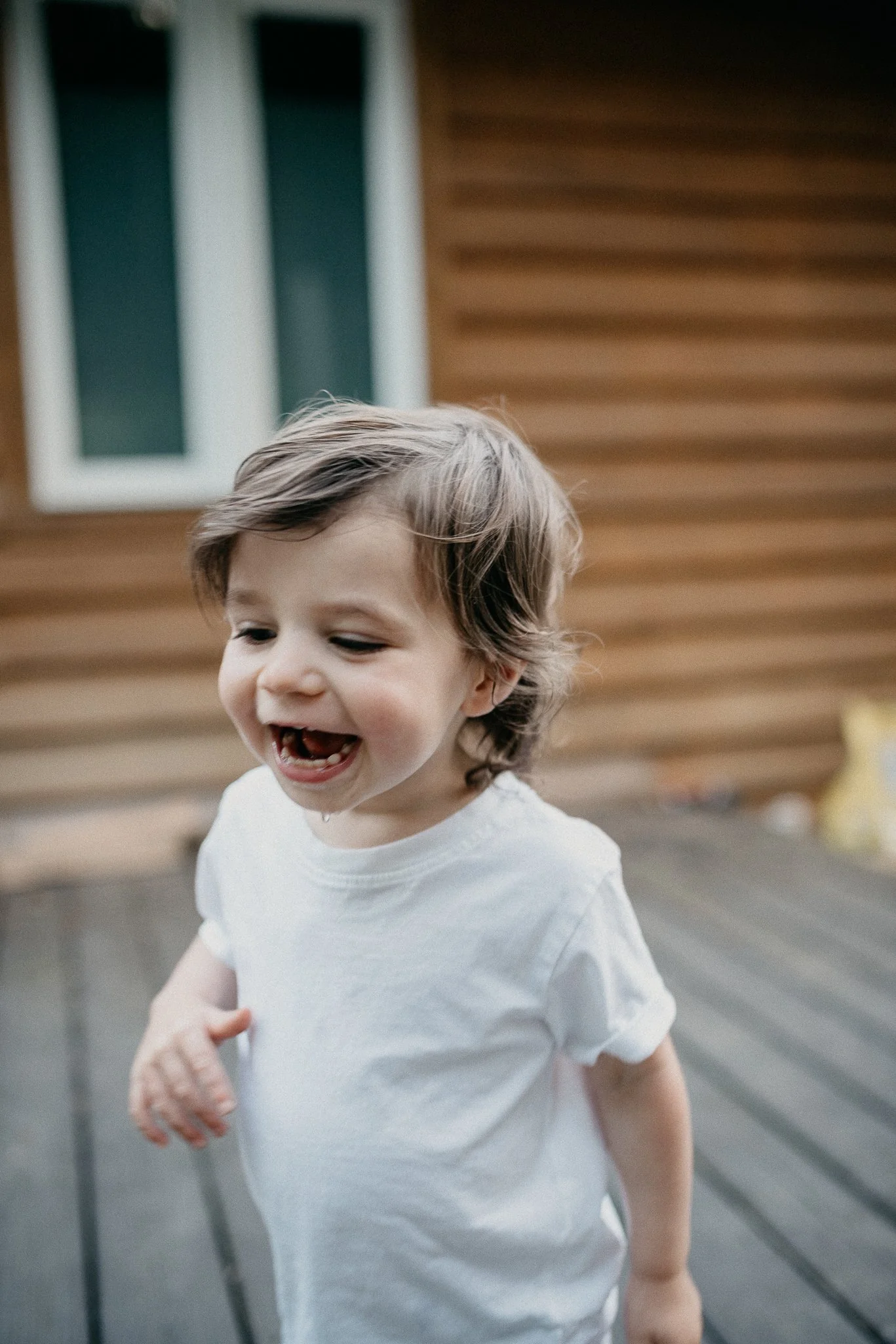 A young boy with light brown hair smiling and running outside near a wooden house with a window.