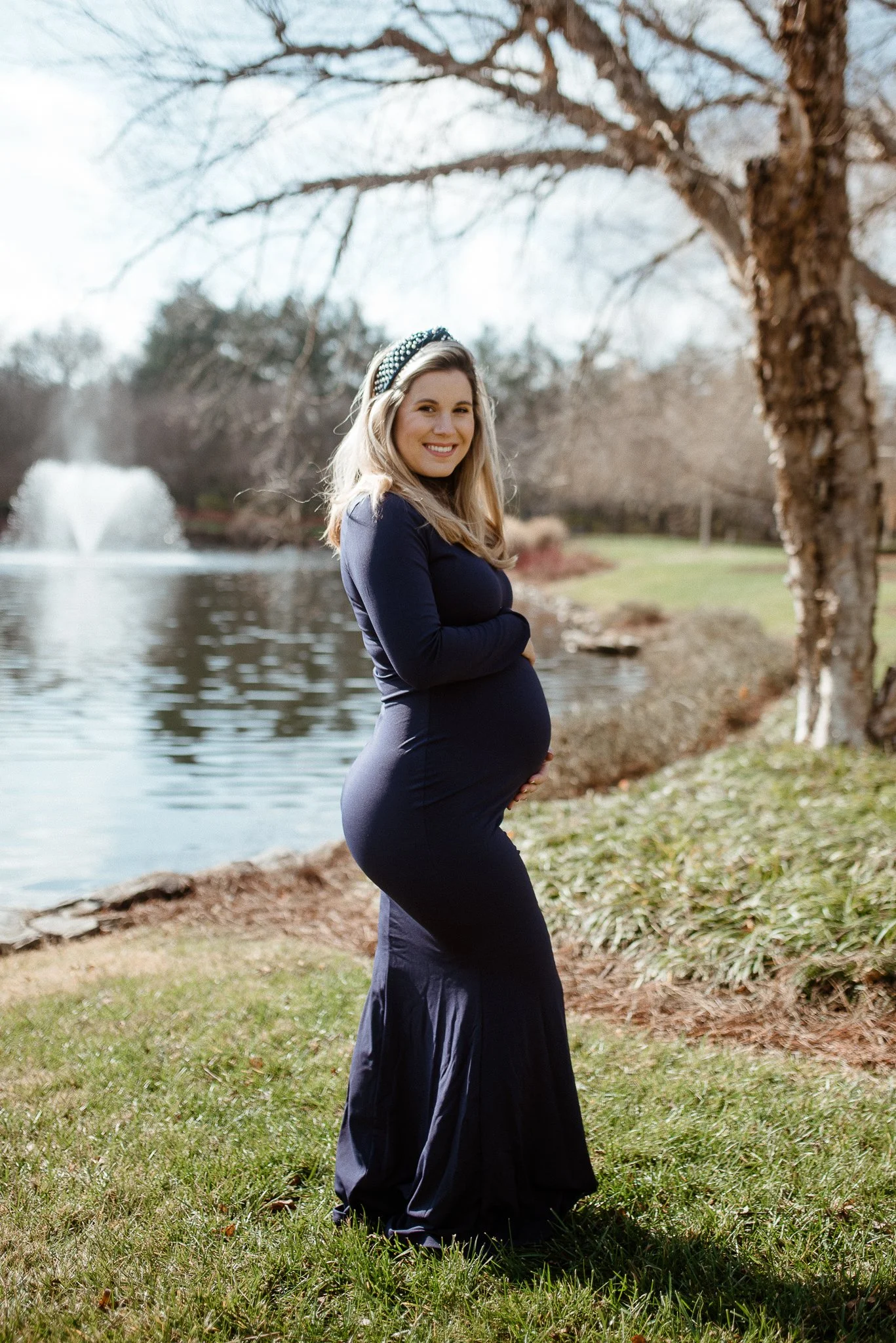 A pregnant woman with blonde hair, wearing a navy blue dress and a beaded headband, standing outdoors by a lake with a fountain, bare trees, and a grassy area in the background, smiling at the camera.
