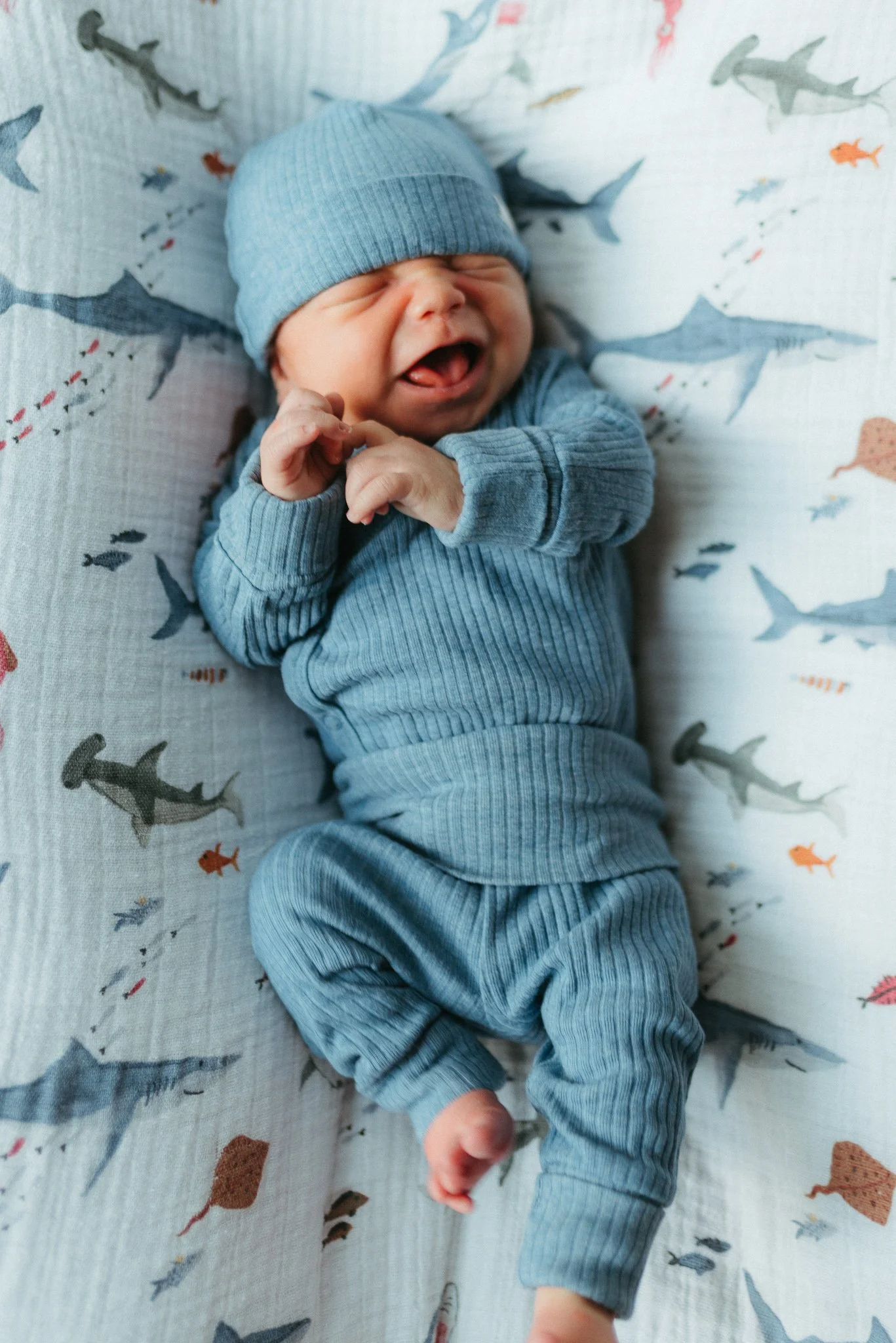 A smiling baby dressed in a blue outfit and a matching blue hat, lying on a bed with a whale and fish print sheet.