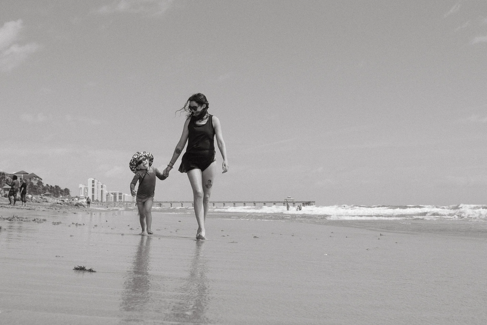 A woman and a young girl holding hands walking along the shoreline at the beach with buildings and a pier in the background.