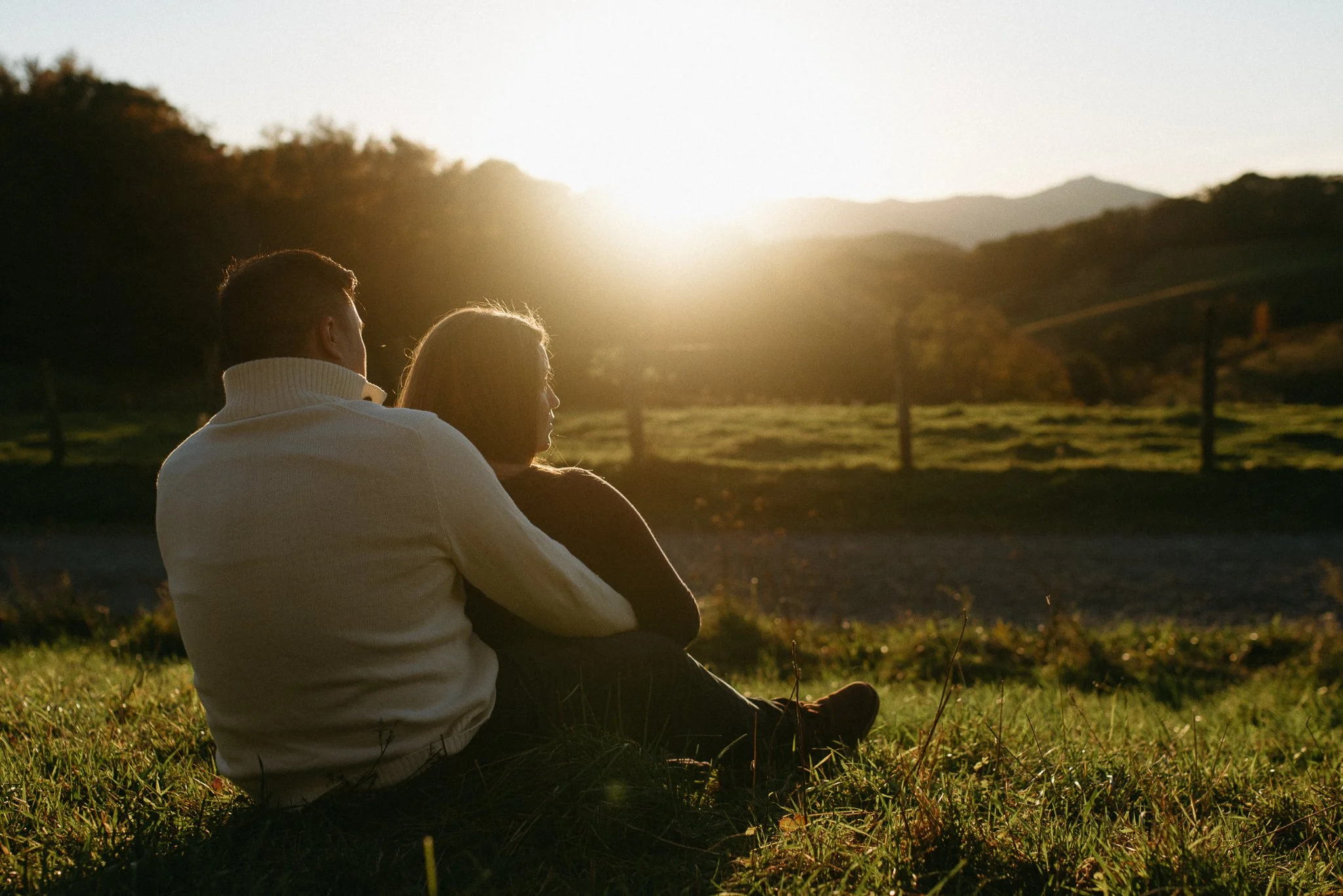 A couple sitting outdoors on the grass during sunset, looking at the horizon with mountains and trees in the background.