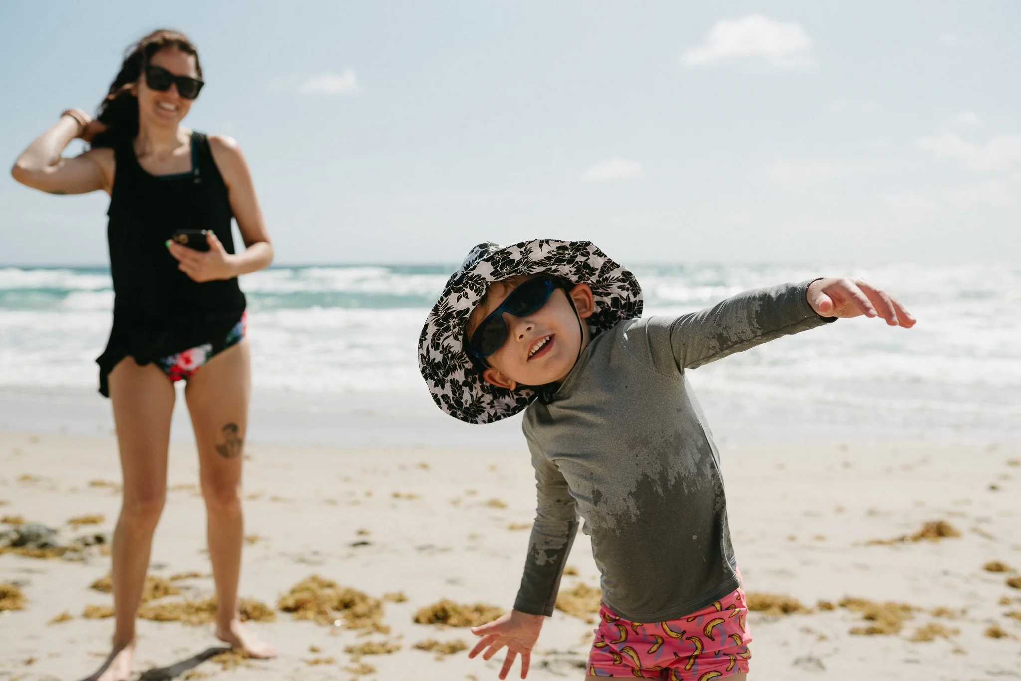 A woman in sunglasses and a black swimsuit with a tattoo on her thigh stands behind a young girl with sunglasses, a sun hat, and pink shorts with bananas, at the beach. The girl is smiling and leaning forward with her hand extended.