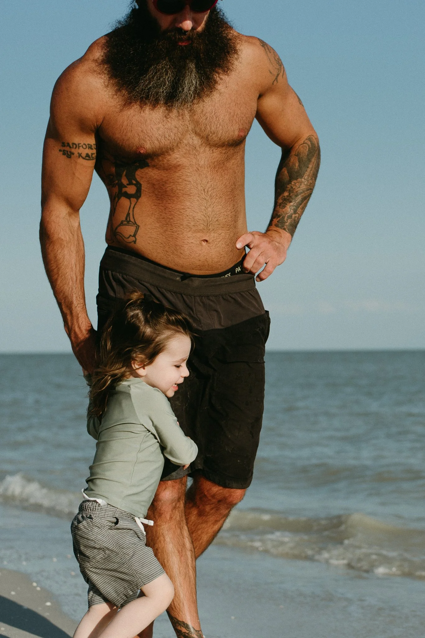 A shirtless man with tattoos on his arm and torso walking on the beach with a young girl, holding her hand. The man has a long beard and dark sunglasses, and the girl is wearing a light green jacket and striped shorts. The ocean and a clear sky are i