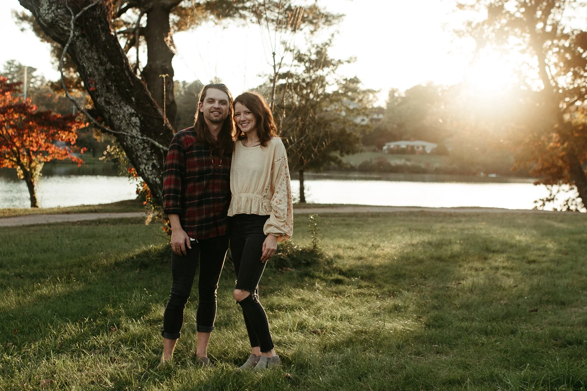 A young man and woman standing together outdoors near a tree by a body of water during sunset, smiling and posing for the photo.