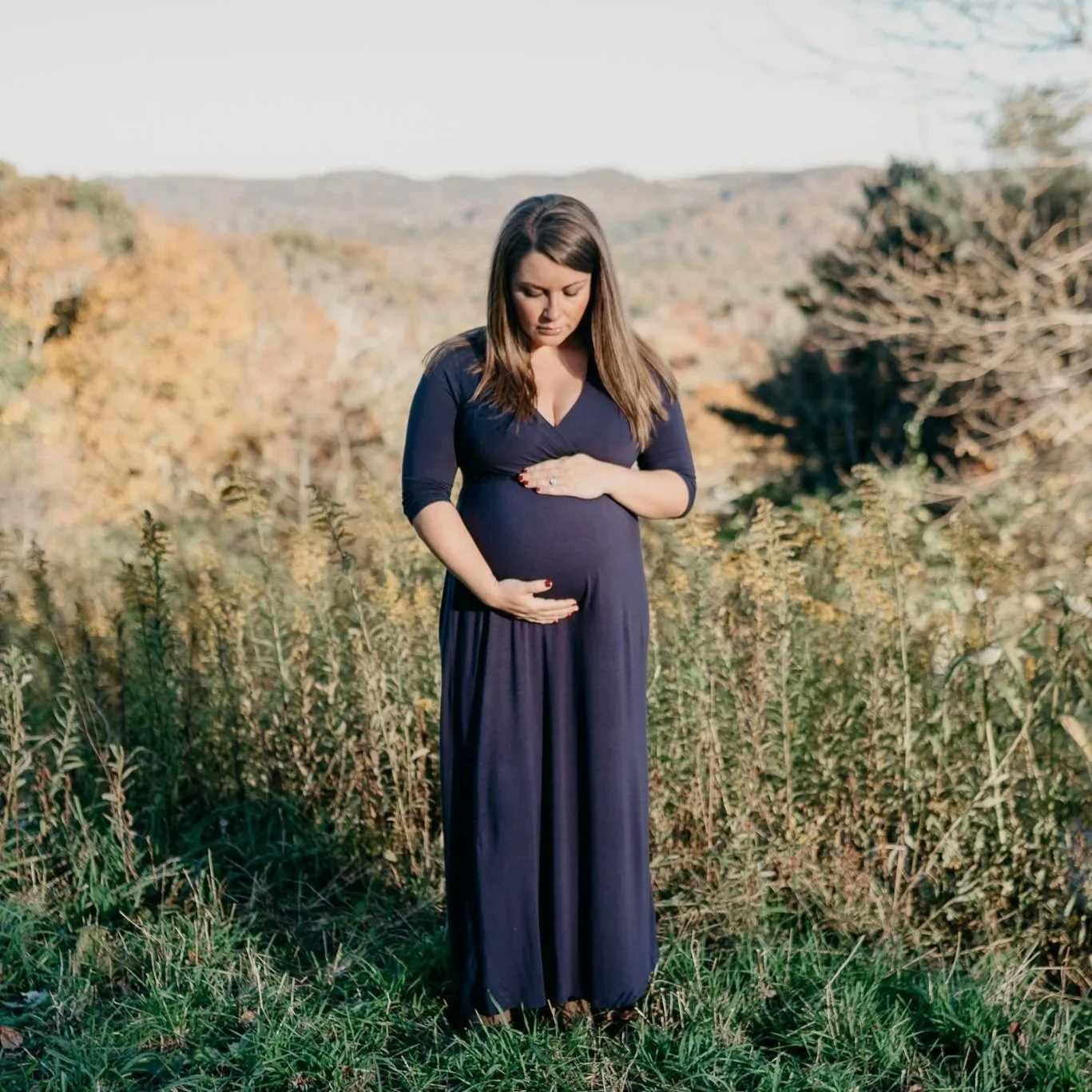 A pregnant woman in a dark blue dress standing in a field with a scenic mountainous background, gently holding her belly with both hands, looking downwards.