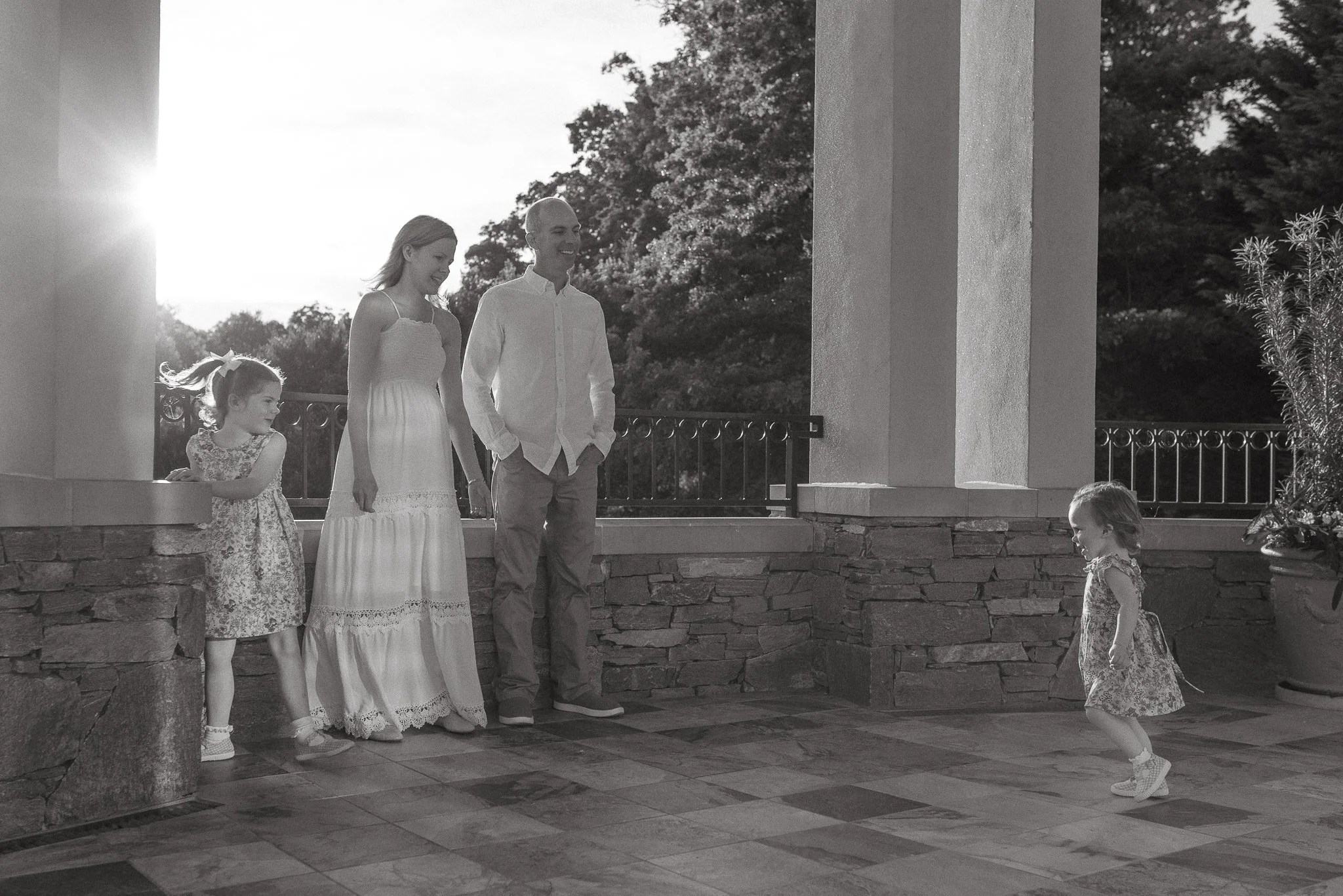 A man and woman holding hands with two young girls, standing on a porch with trees in the background, during sunset.