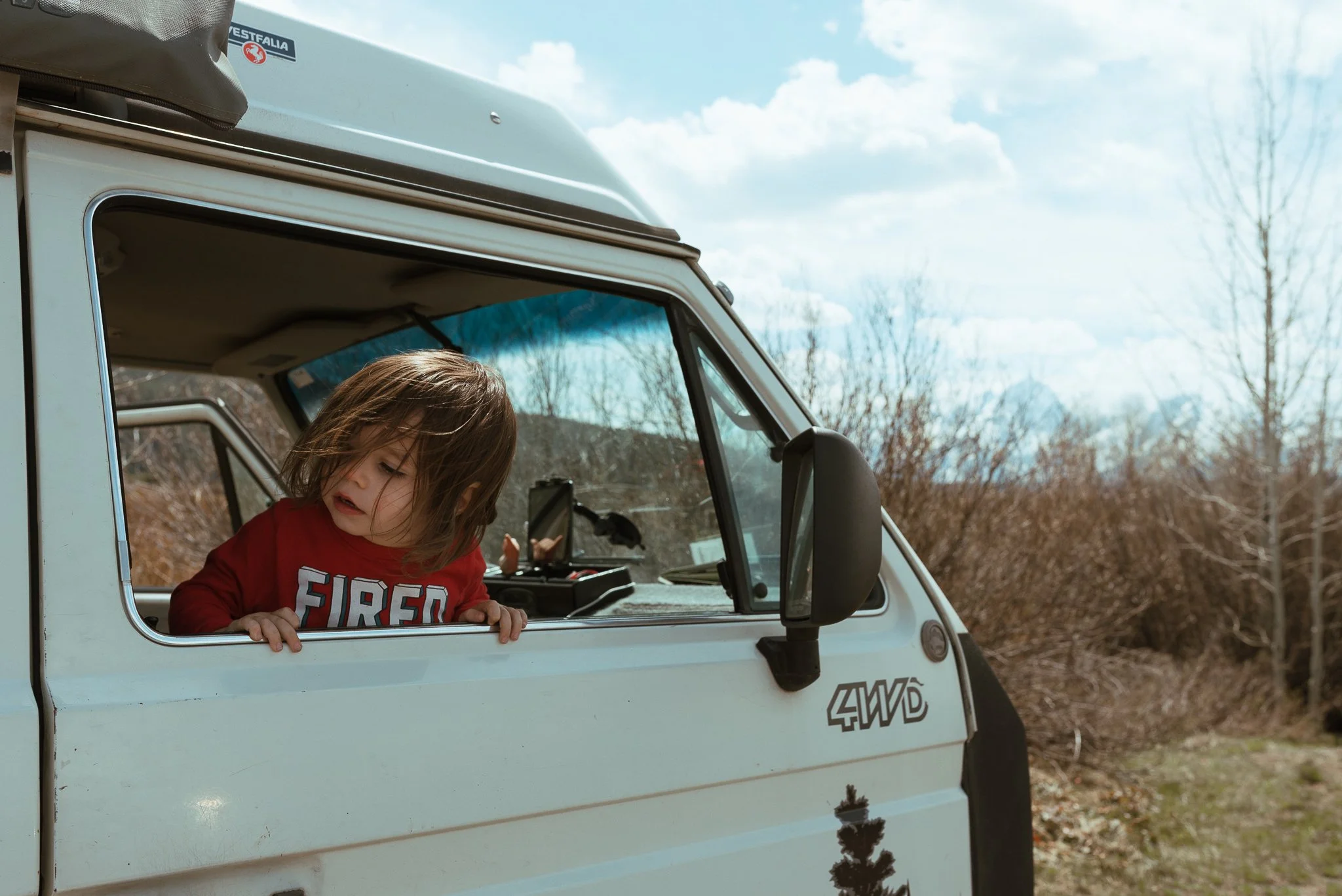 A young child with long brown hair, wearing a red Fire Department shirt, is leaning out of the window of a white off-road vehicle with a child window. The vehicle is parked outdoors on a dirt path in a rural area with trees and mountains in the backg