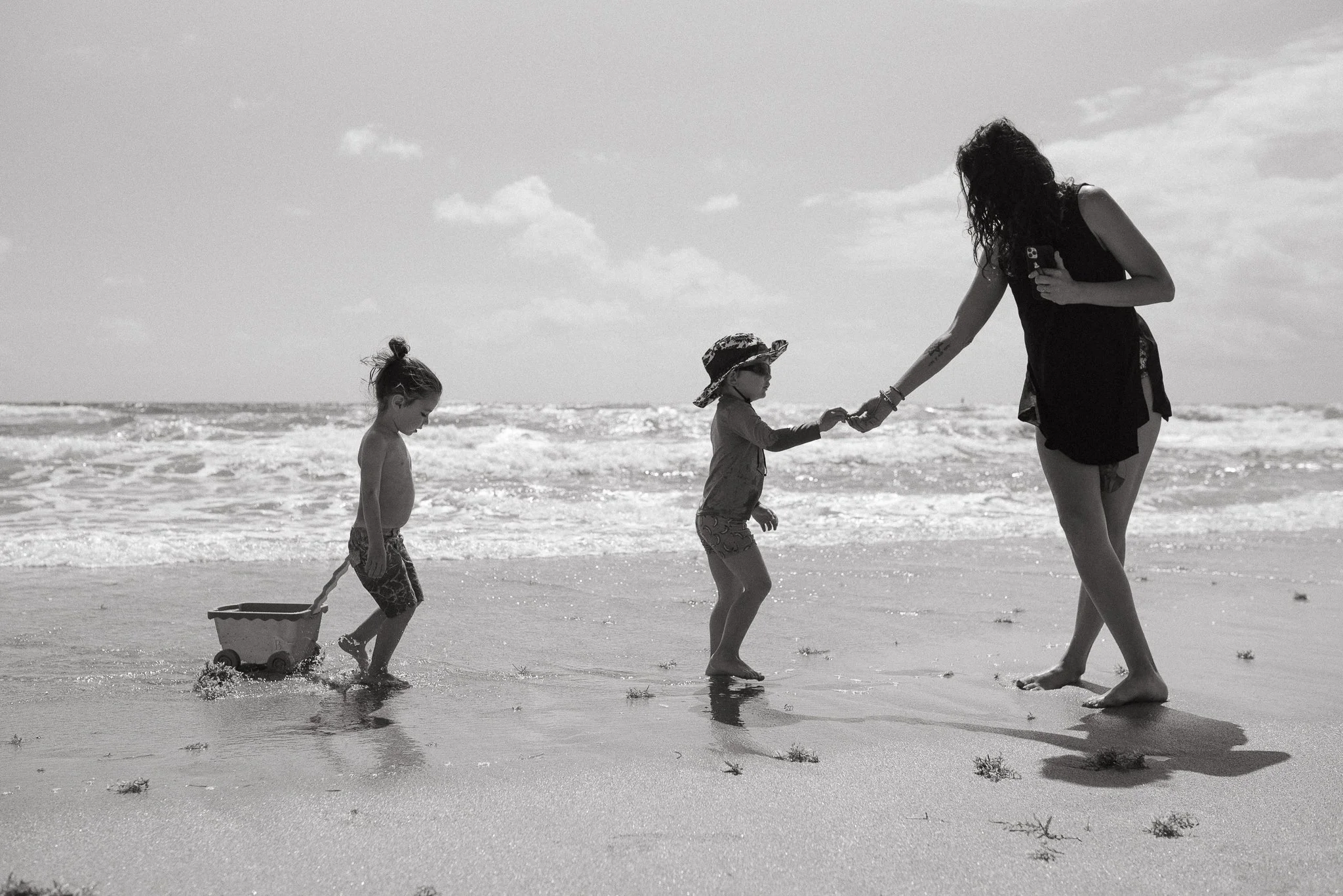 Black-and-white photo of a woman and three children at the beach. The woman is holding hands with one child, while another child pulls a wagon. The children are wearing hats, and the woman is wearing a dress. The ocean and sky are visible in the back