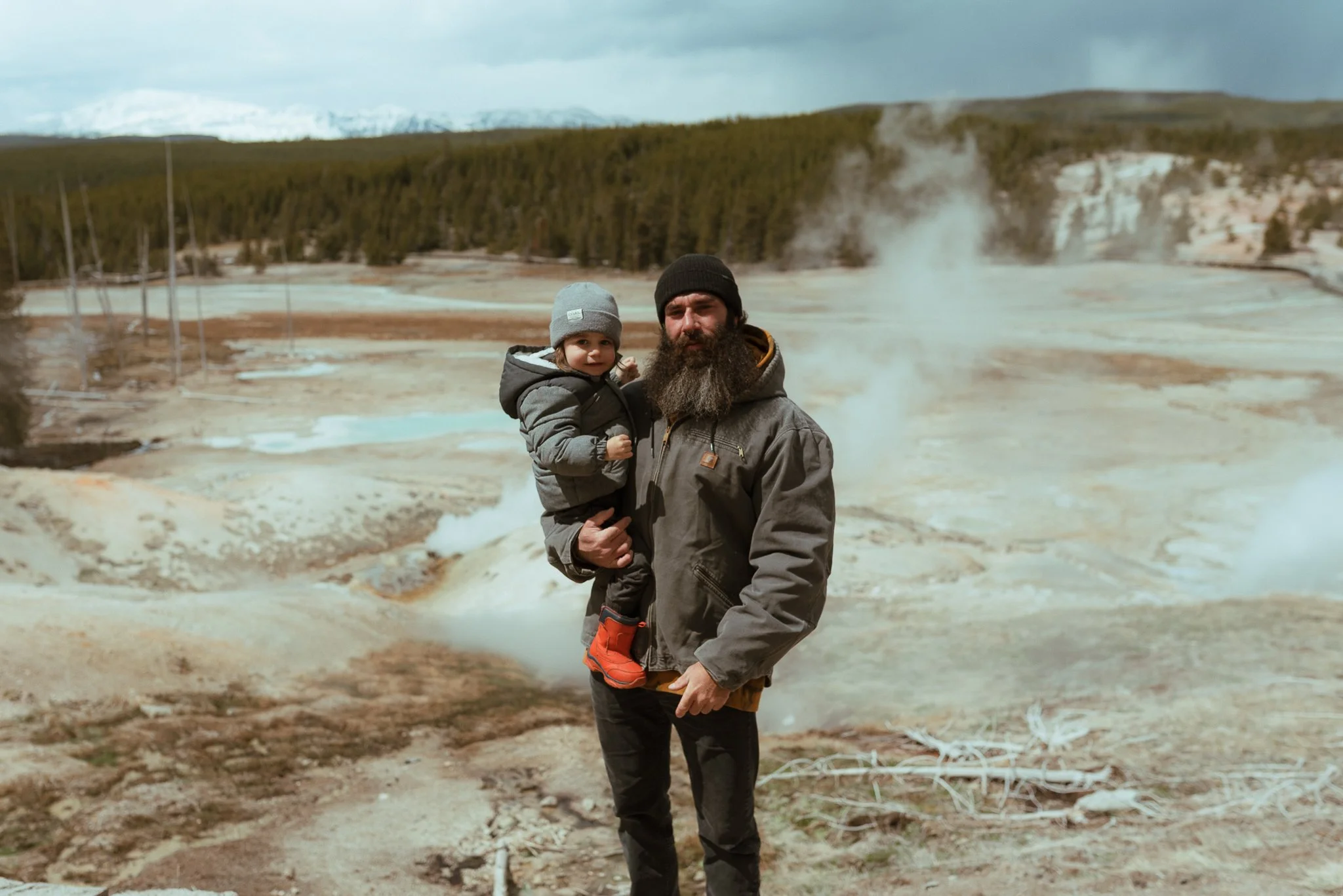 A man with a beard and a child in a gray hat and jacket standing in front of a geothermal area with steaming vents, barren land, and a forested hillside in the background.