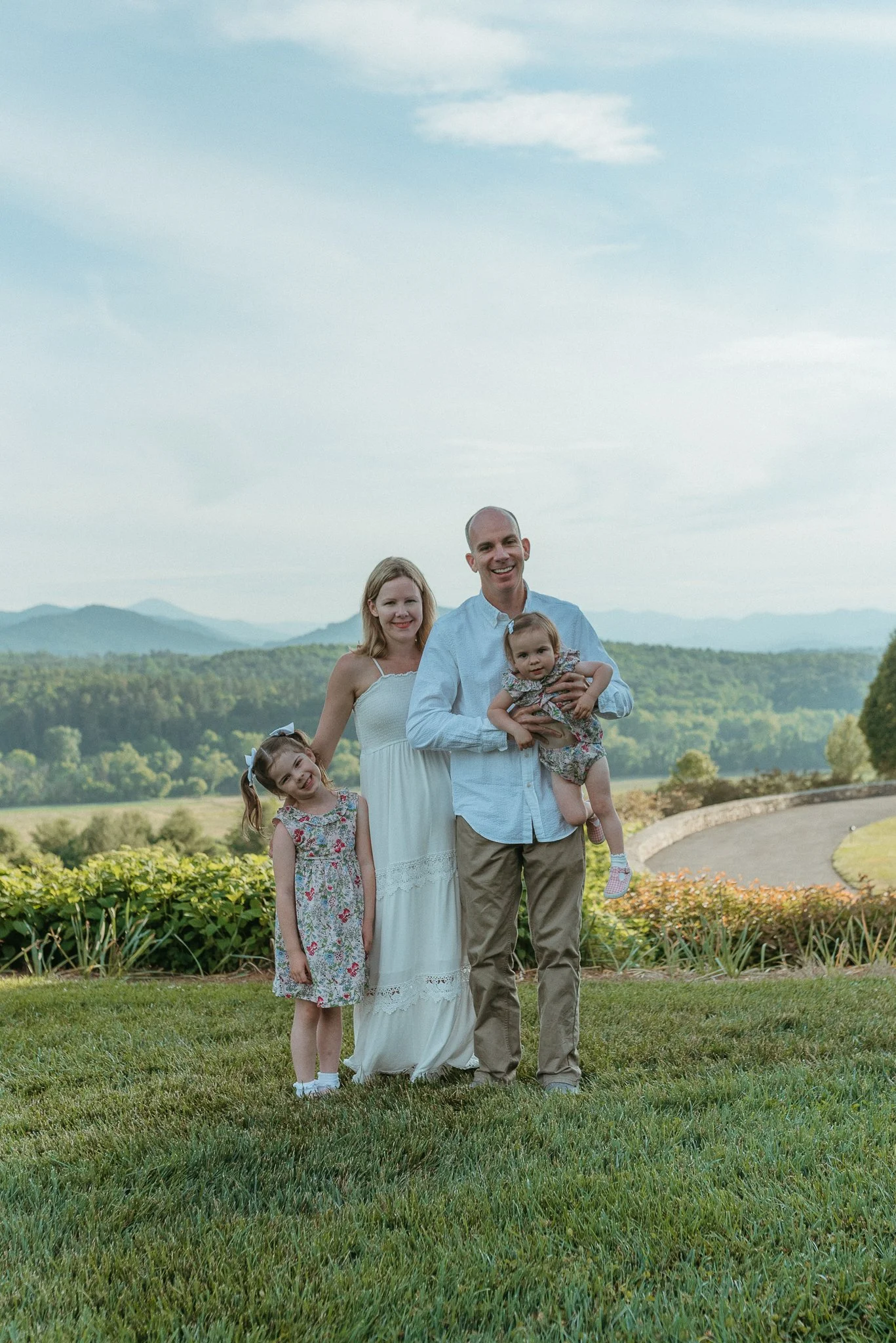 A happy family of five standing outdoors in a park with mountains in the background, smiling at the camera. The family includes a man, a woman, and three young girls, with the girls wearing floral dresses.