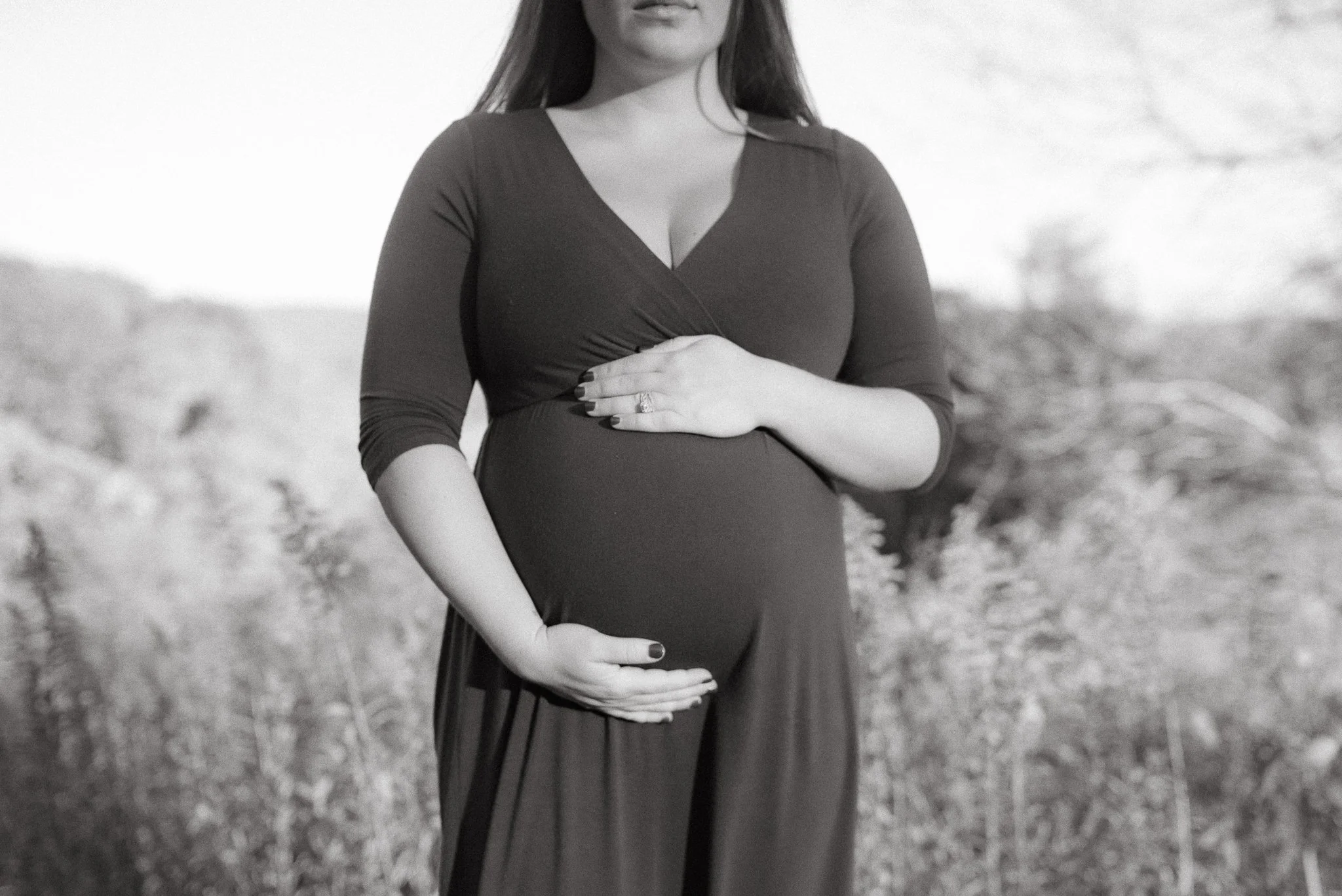 A woman in a dark dress with three-quarter sleeves standing outdoors, holding her stomach. The photo is in black and white.