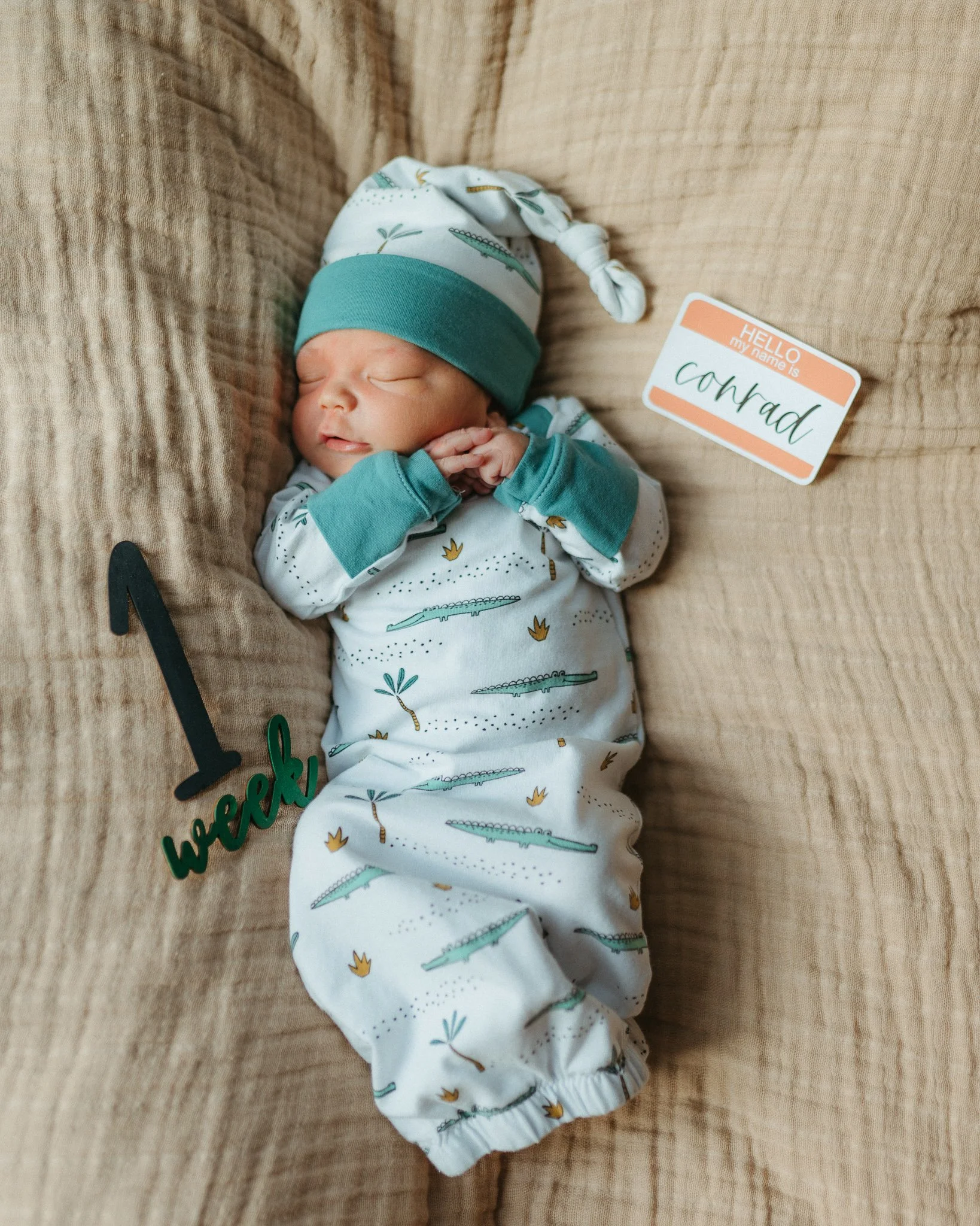 Newborn baby sleeping on a beige textured blanket, wearing pajamas with crocodile and plant patterns, and a white and teal hat. There is a 'hello my name is' badge with the name 'Conrad' placed next to the baby.