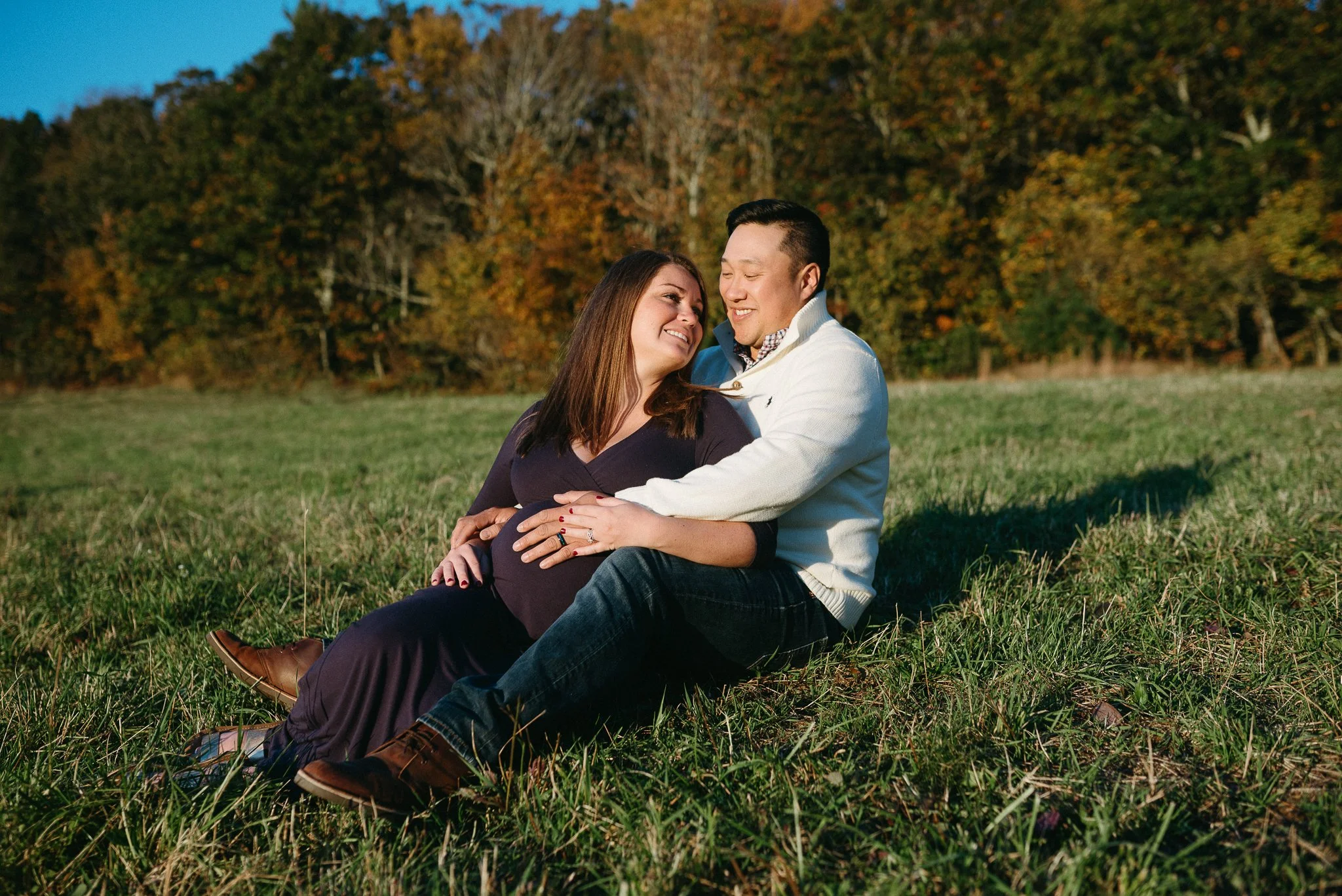 A smiling couple sitting on grass in a park with trees showing fall foliage, sharing a close and affectionate moment.