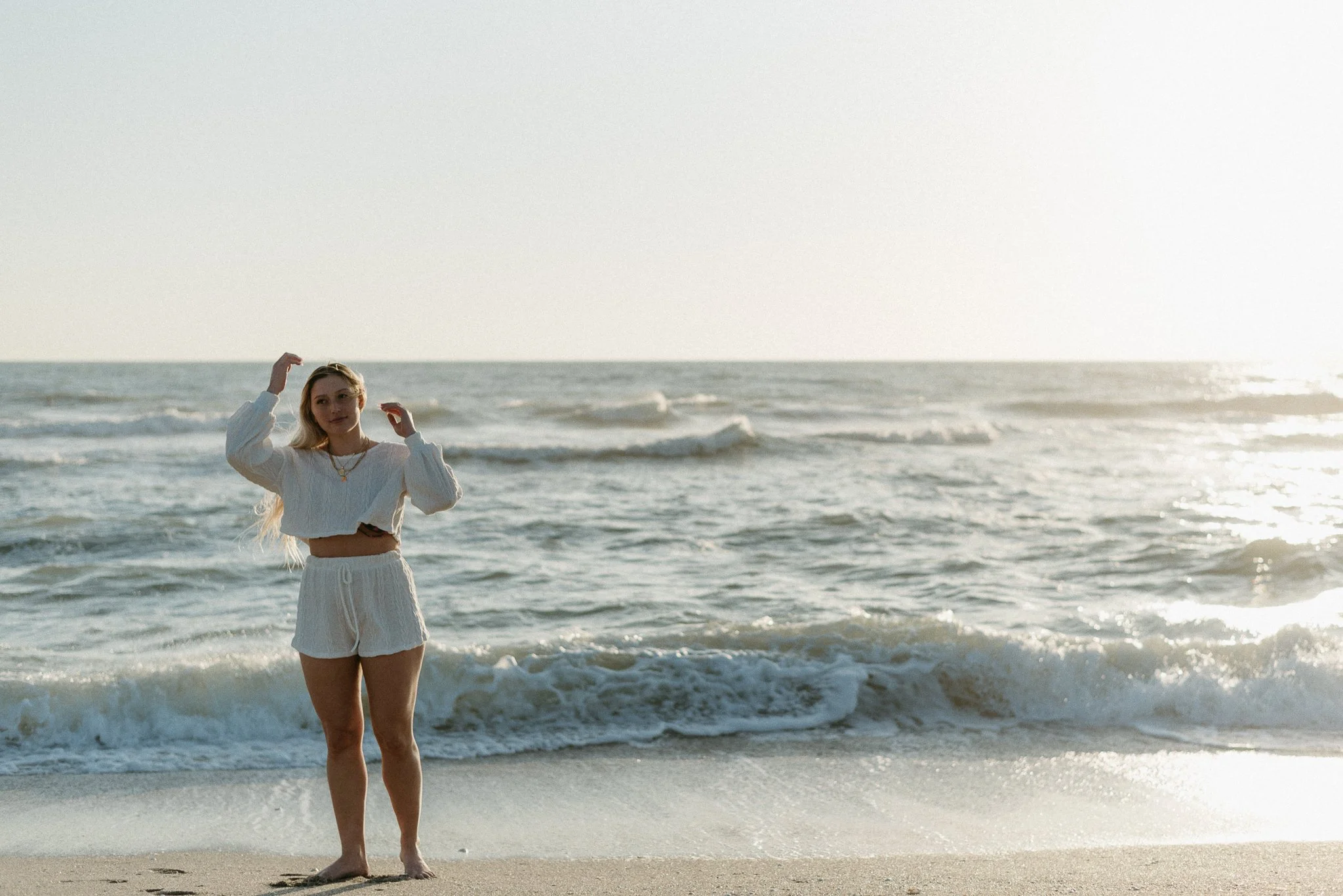 A woman with long blonde hair in a white sweater and shorts standing on the beach near the ocean during sunset.