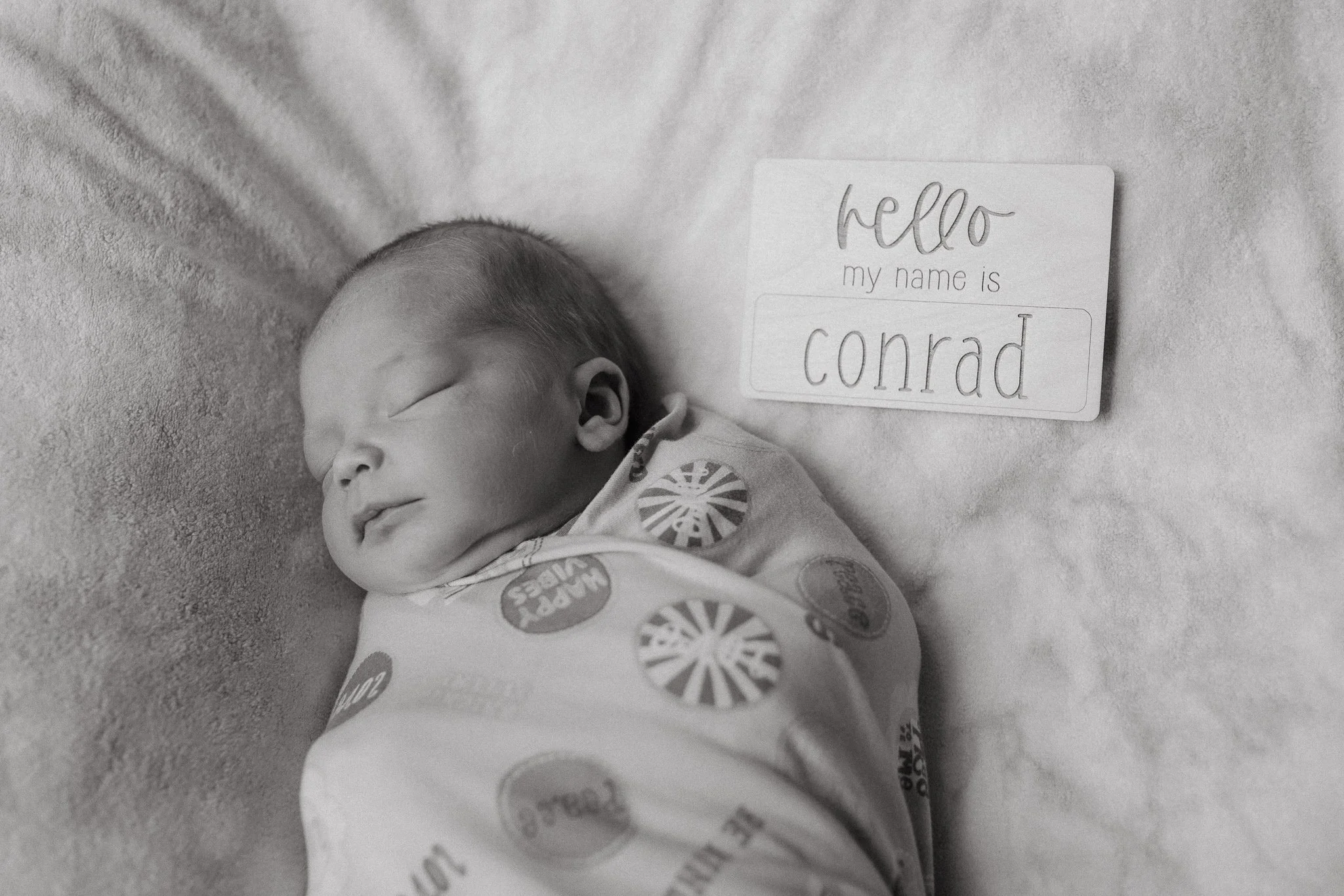 Black and white photo of a sleeping baby with a note beside them that reads 'hello my name is conrad.' The baby is lying on a soft surface, wearing a onesie with circular patterns.