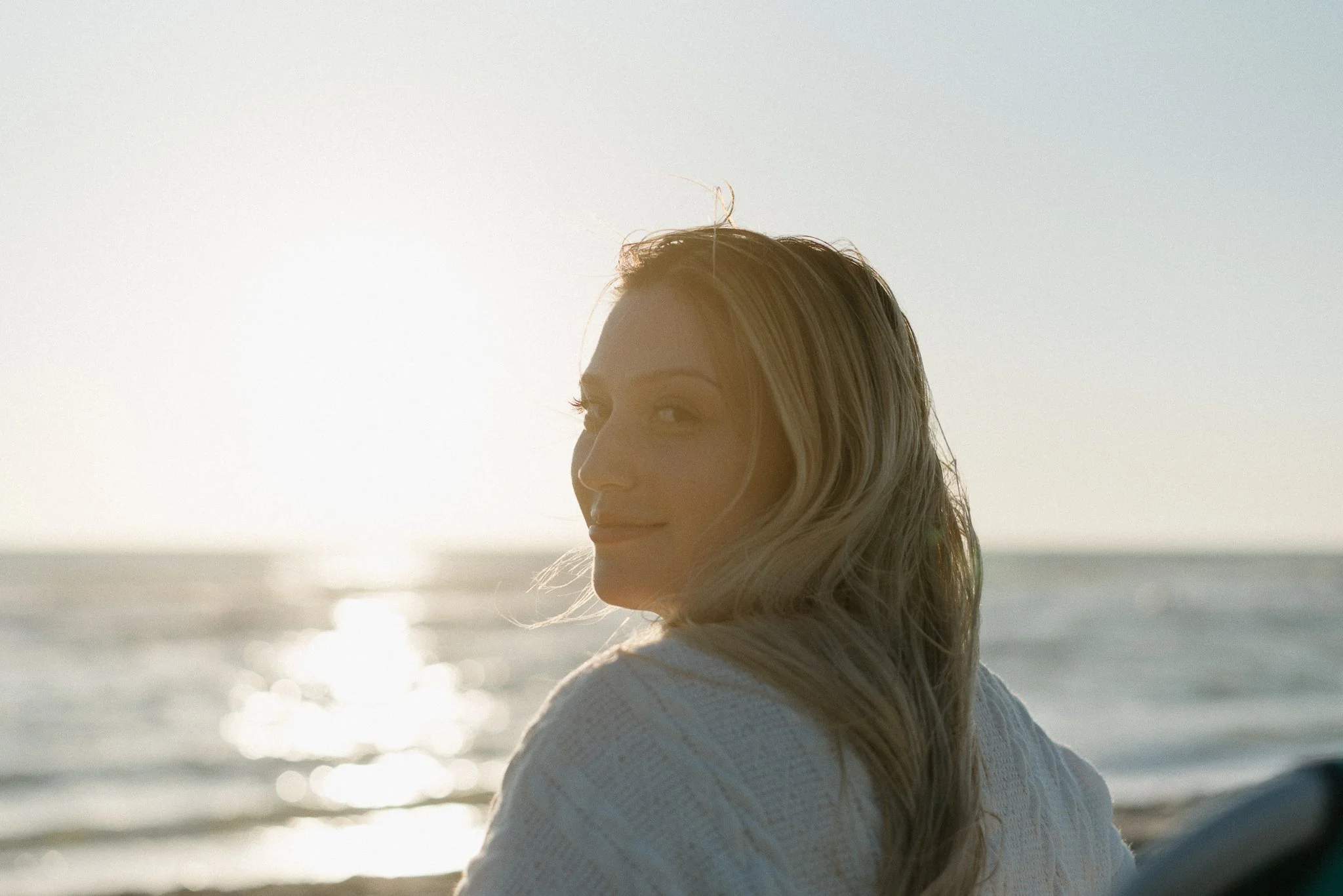 A woman with light-colored hair looking at the ocean at sunset or sunrise, with sunlight reflecting off the water.