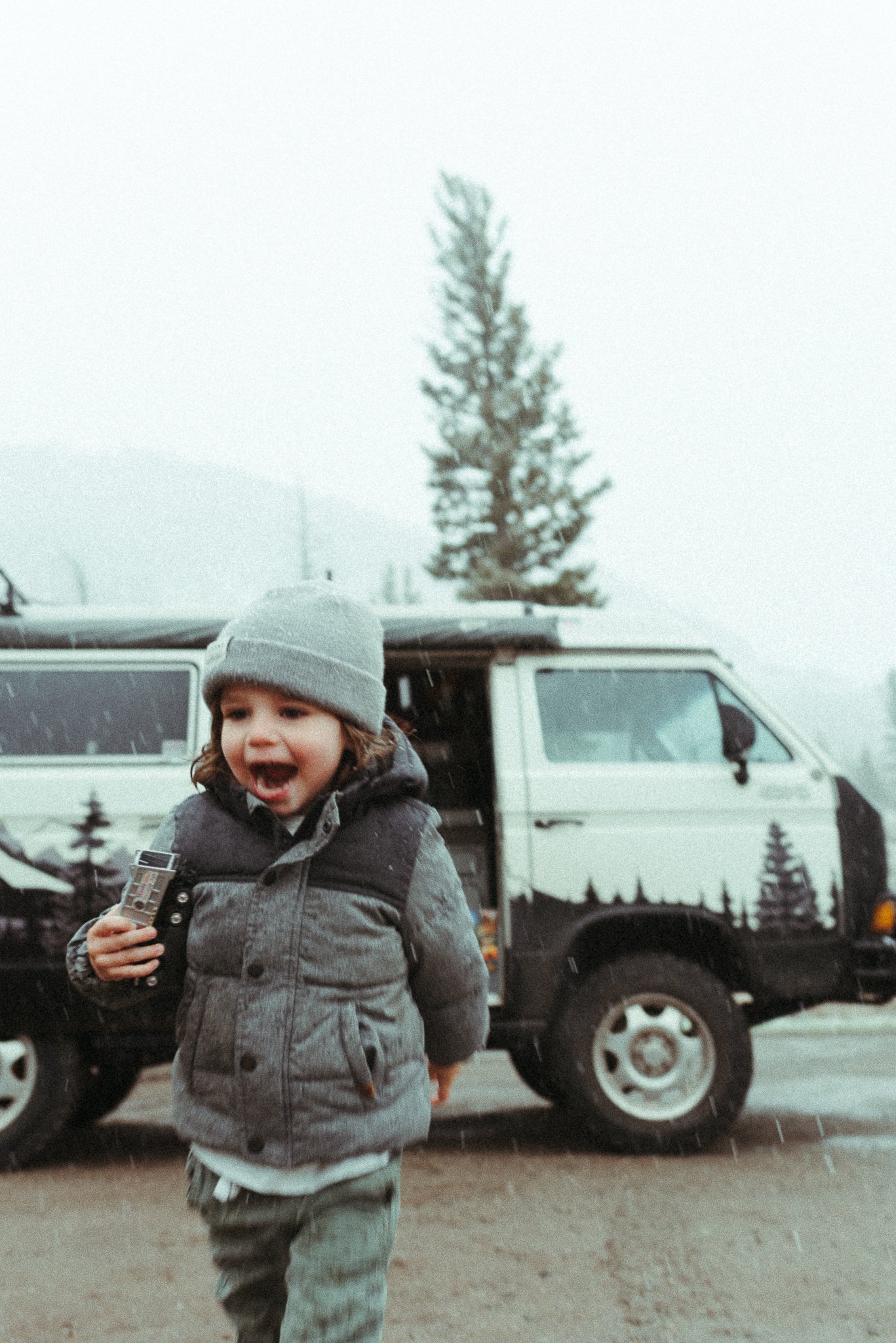 Child with curly hair wearing a gray jacket and hat, smiling and holding a toy, standing in front of a white off-road vehicle during winter.