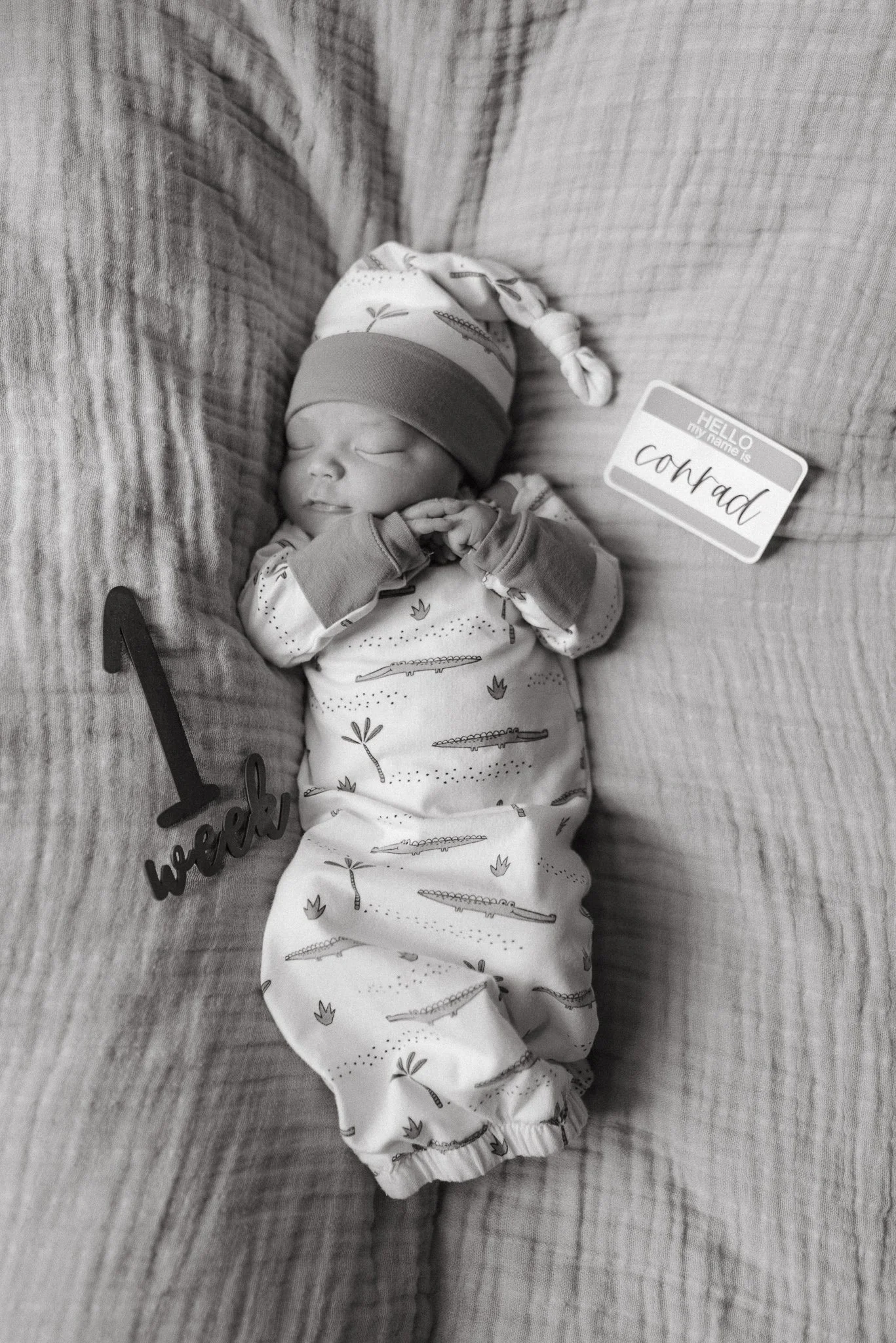 A sleeping newborn baby wearing a hat and a printed onesie, lying on a textured surface with a 'hello new' name tag nearby.