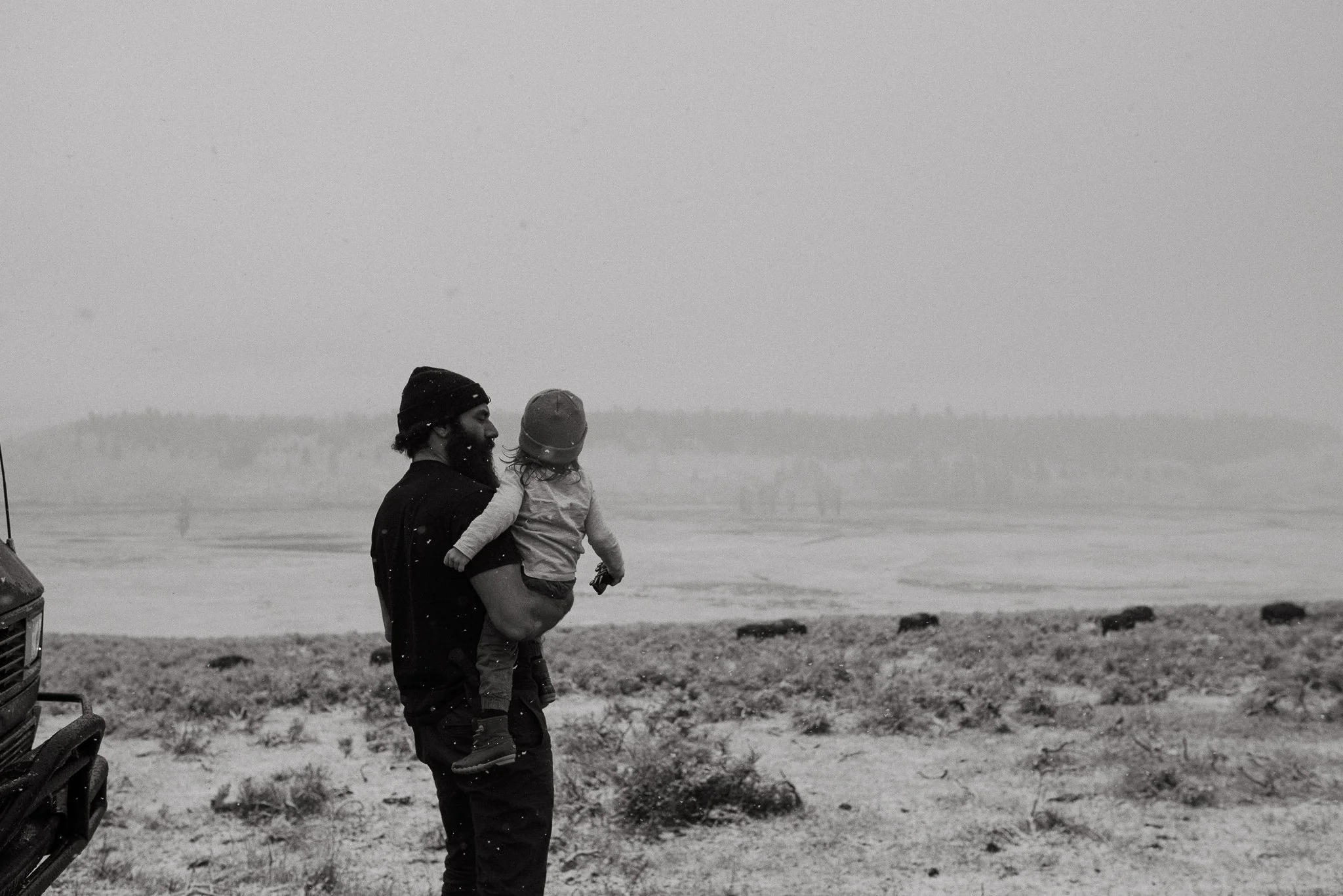 A man holding a child on a beach during snowy weather, both wearing warm clothing and beanies, with a foggy, overcast sky in the background.
