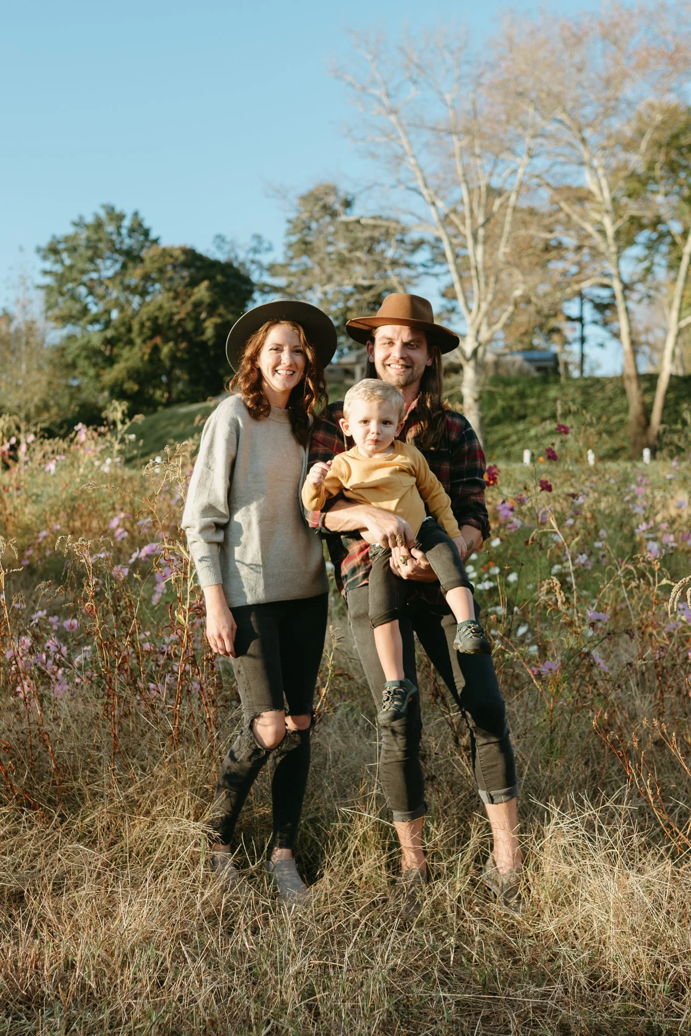 A family of three standing outdoors in a field of tall grass and wildflowers, smiling at the camera. The woman wears a gray sweater and a black wide-brimmed hat, the man wears a plaid shirt and a brown hat, and the child, being held by the man, wears