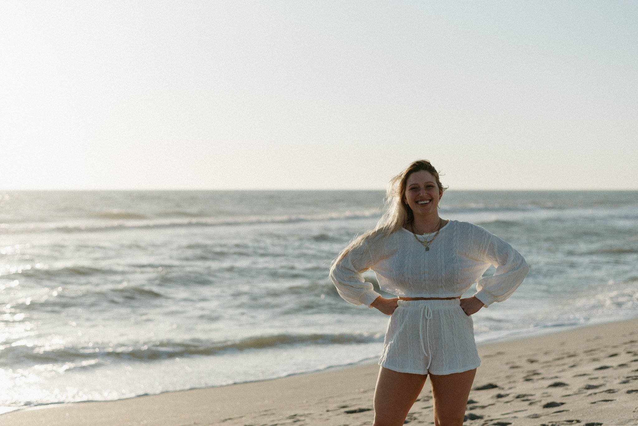 A woman standing on a beach, smiling with hands on hips, wearing a white sweater and shorts, with ocean waves in the background.