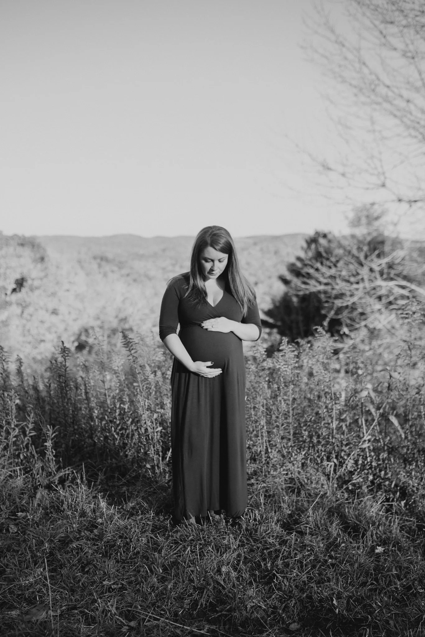 A pregnant woman in a long dress stands in a field of tall grass, holding her belly with both hands, with mountains and leafless trees in the background, in black and white.