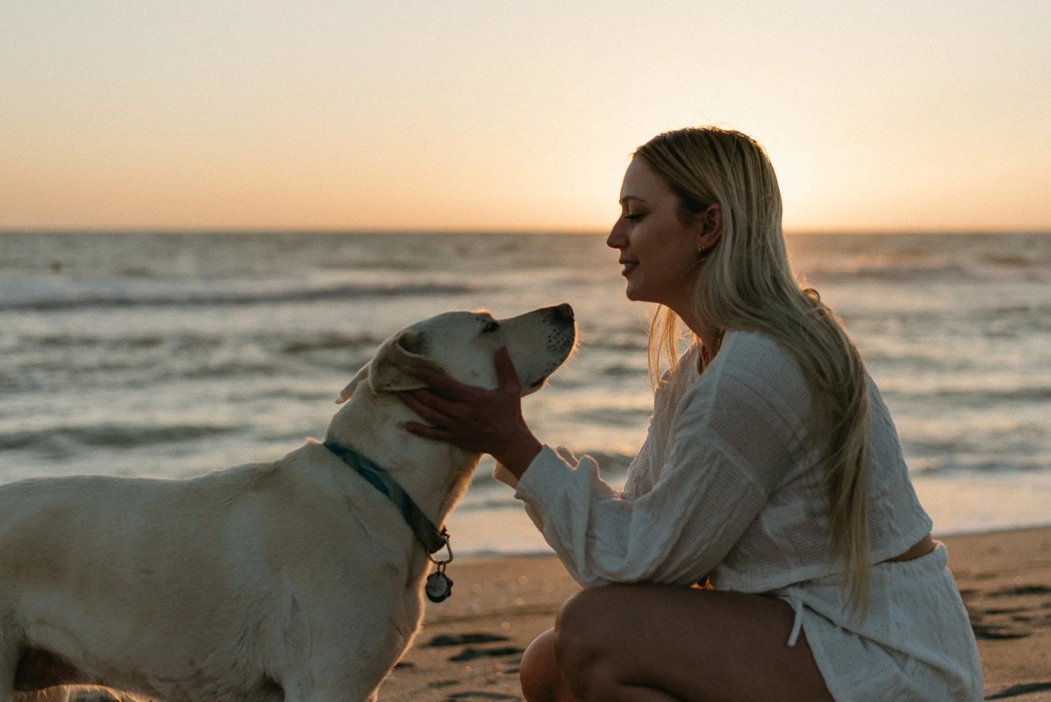 A woman with long blonde hair and a white sweater kneeling on the beach, face close to a yellow Labrador retriever dog, during sunset.