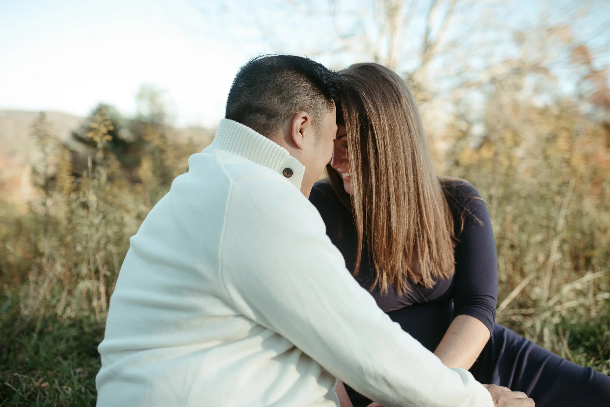 A man and woman sitting outdoors, close together, sharing a moment of affection in a natural setting with trees in the background during fall.