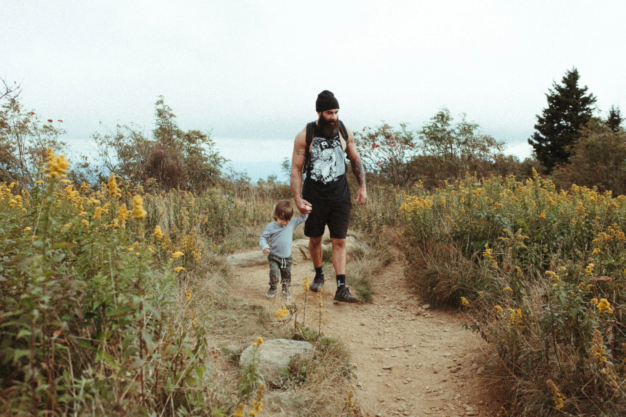 A man with a beard, wearing a black beanie, black shirt with a graphic design, and black shorts, walks with a young child on a dirt trail through a field of yellow wildflowers and shrubs, with tall trees in the background under an overcast sky.