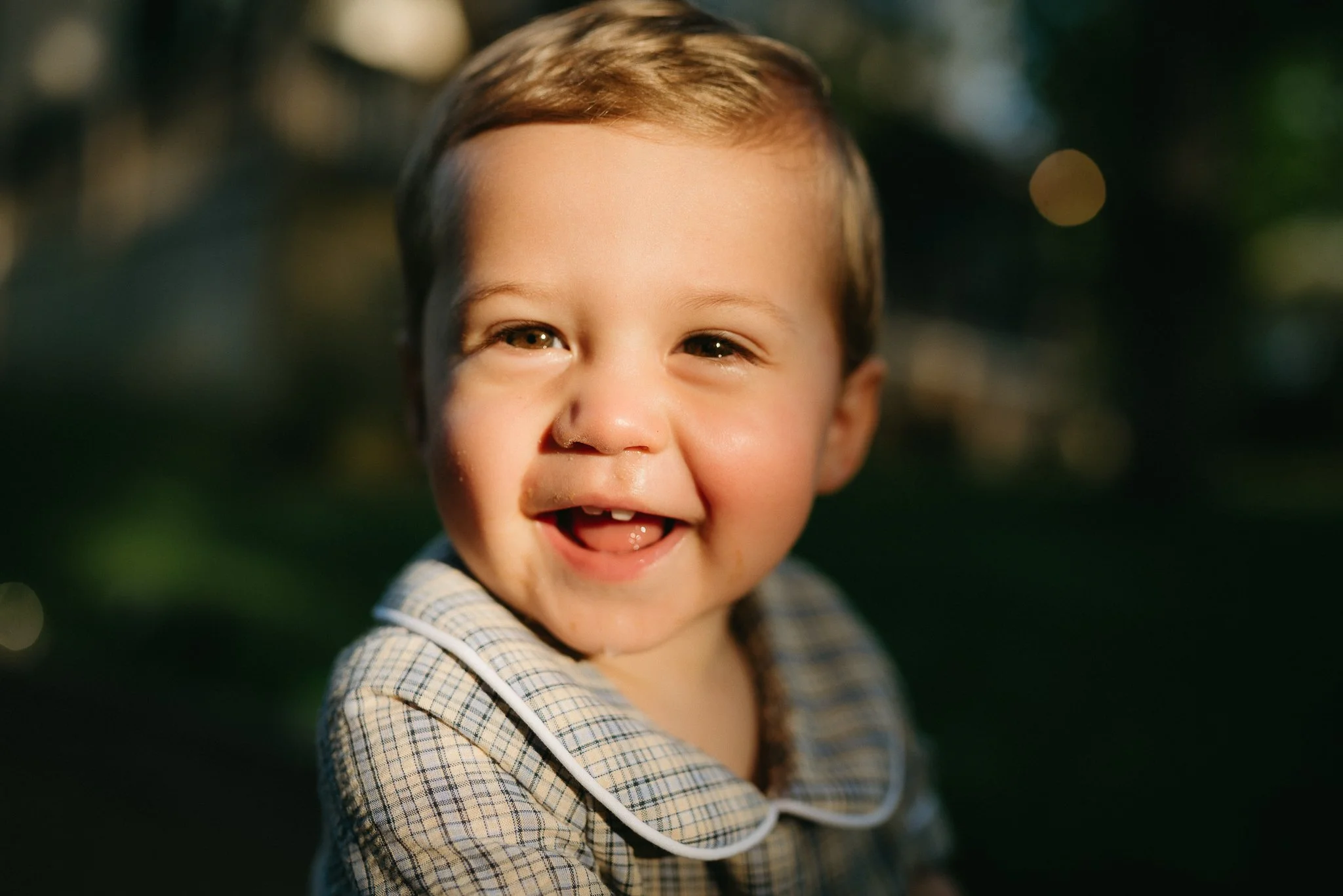 Close-up of a smiling young child with brown hair, wearing a plaid shirt, outdoors during golden hour.