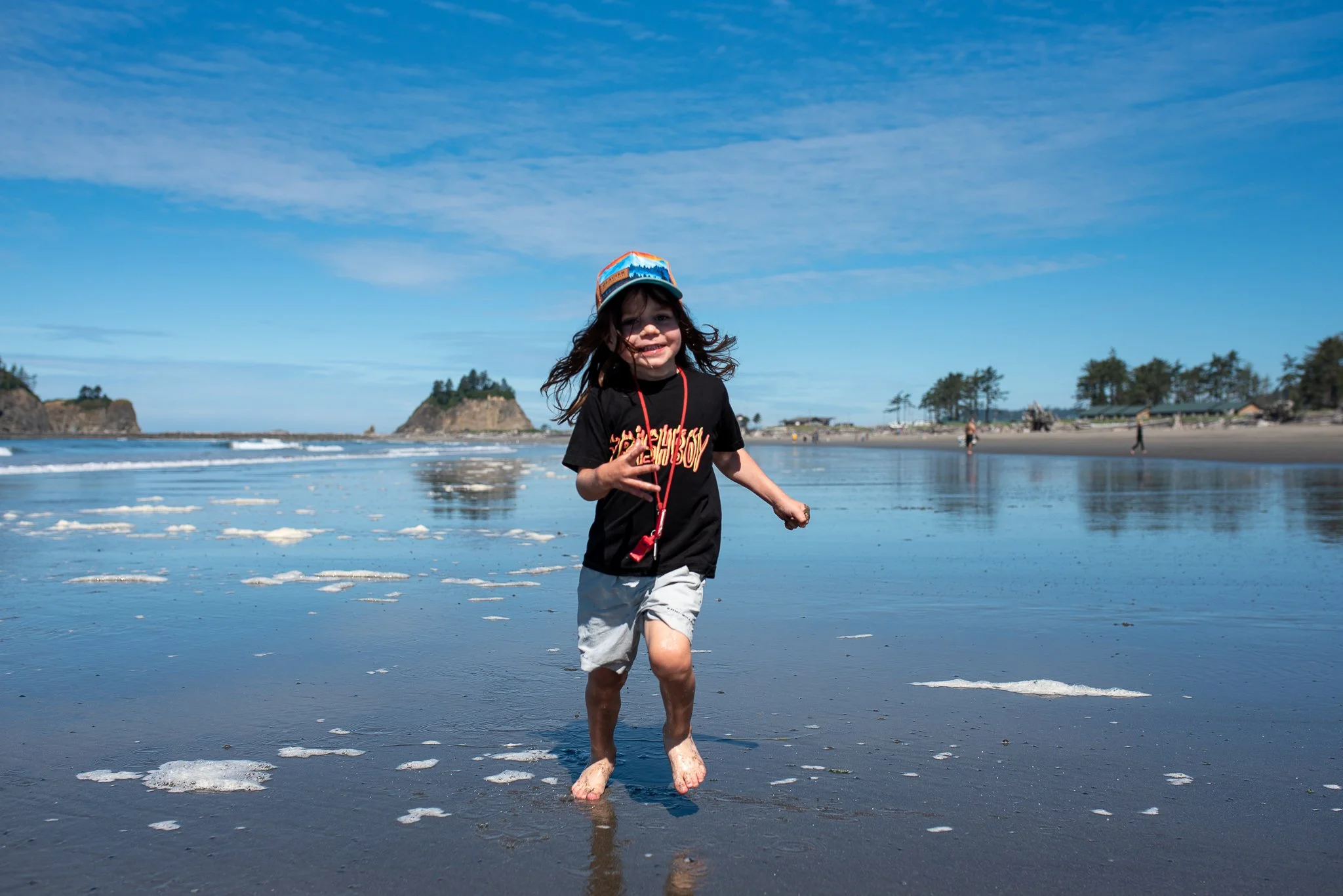 Child running on the beach with footprints in shallow water, wearing a cap, black t-shirt, and shorts, with a bright sky and distant shoreline in the background.