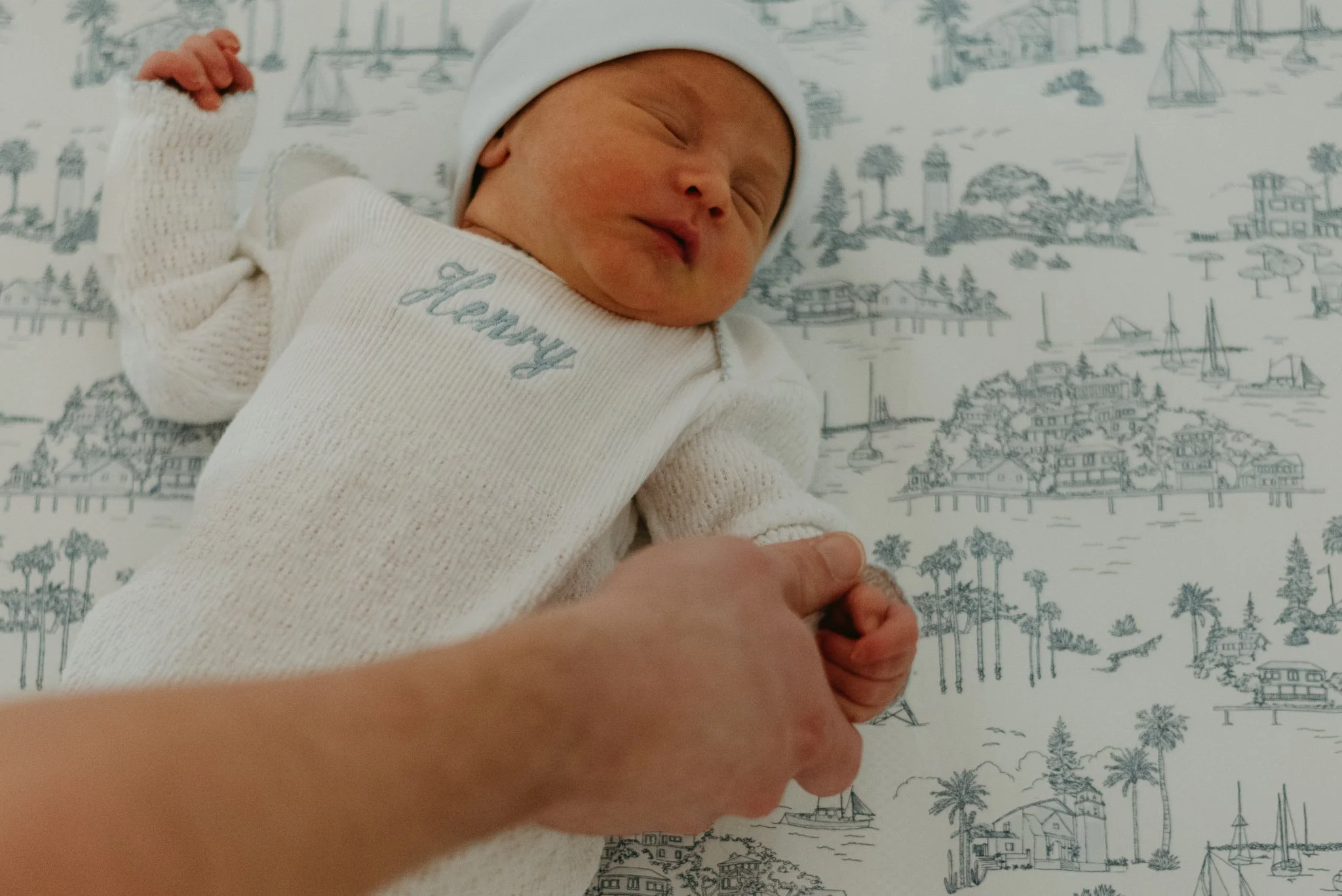 Close-up of a newborn baby lying on a patterned surface, wearing a white hat and sweater, smiling with closed eyes, with child's hand gently holding an adult's finger.