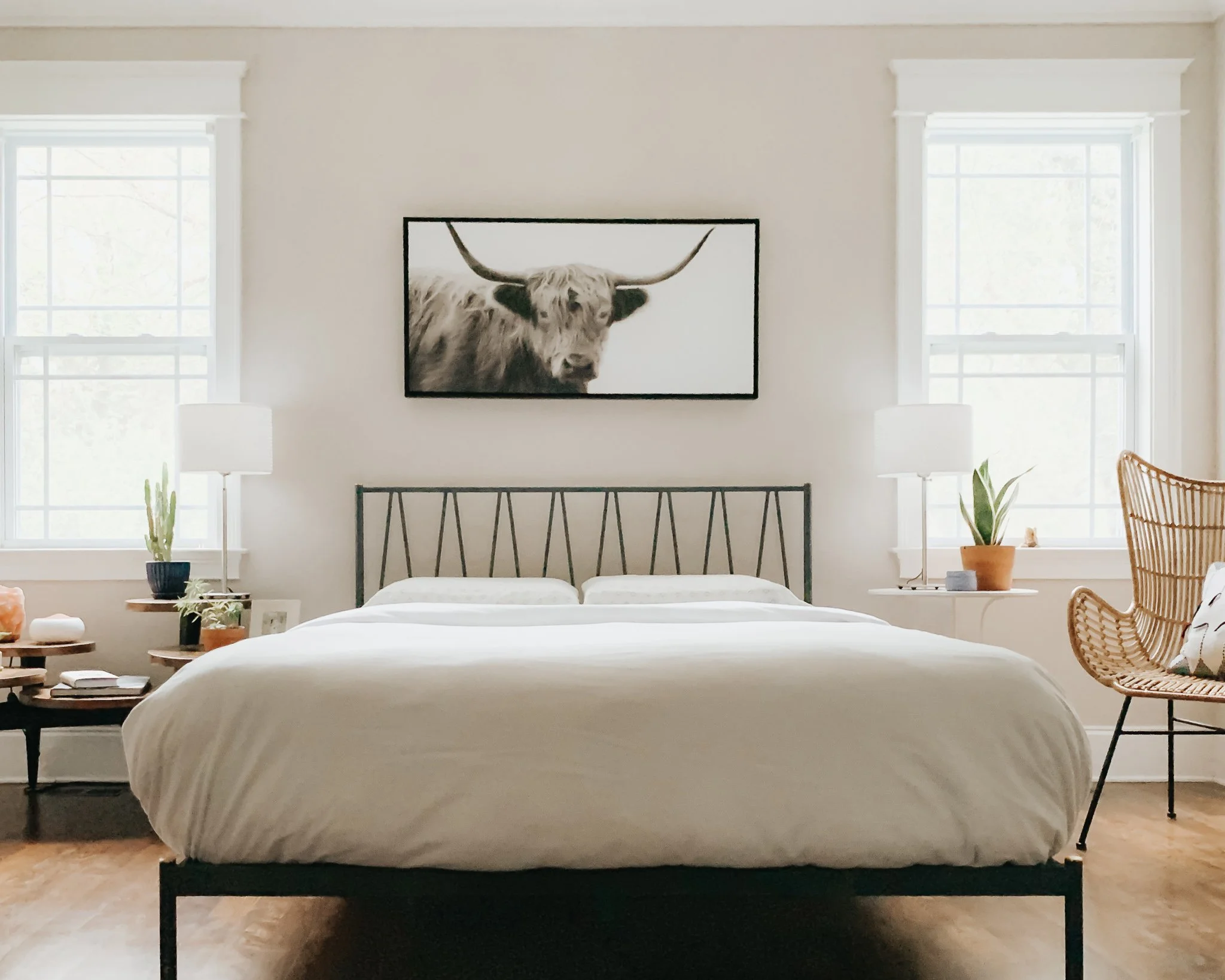 A bedroom with two beds having black metal frames and white bedding, two large windows with white trim, two white nightstands with lamps, and a black-and-white highland cow artwork on the wall above the beds.