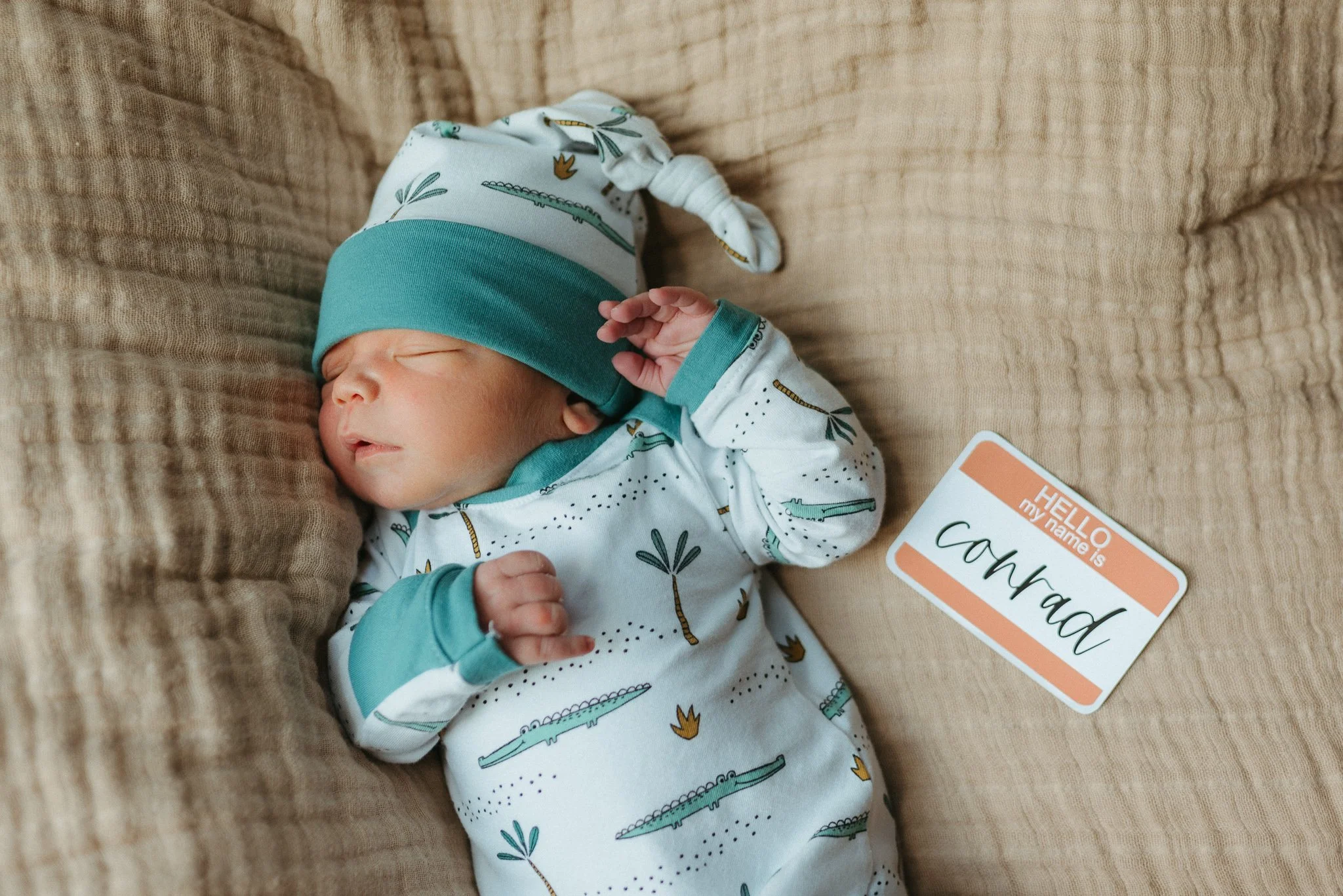 A sleeping baby wearing a patterned onesie and a teal and white hat, lying on a beige textured couch. The baby has a name tag that says 'Cavah', and part of a card reading 'HELLO my name is' is visible near the baby's face.