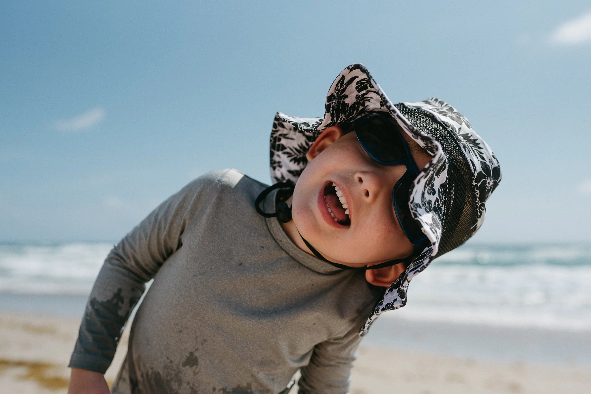 A young boy smiling while playing on the beach. He is wearing sunglasses, a wide-brimmed hat with a floral pattern, and a long-sleeved gray shirt. The background shows the sky and the ocean.