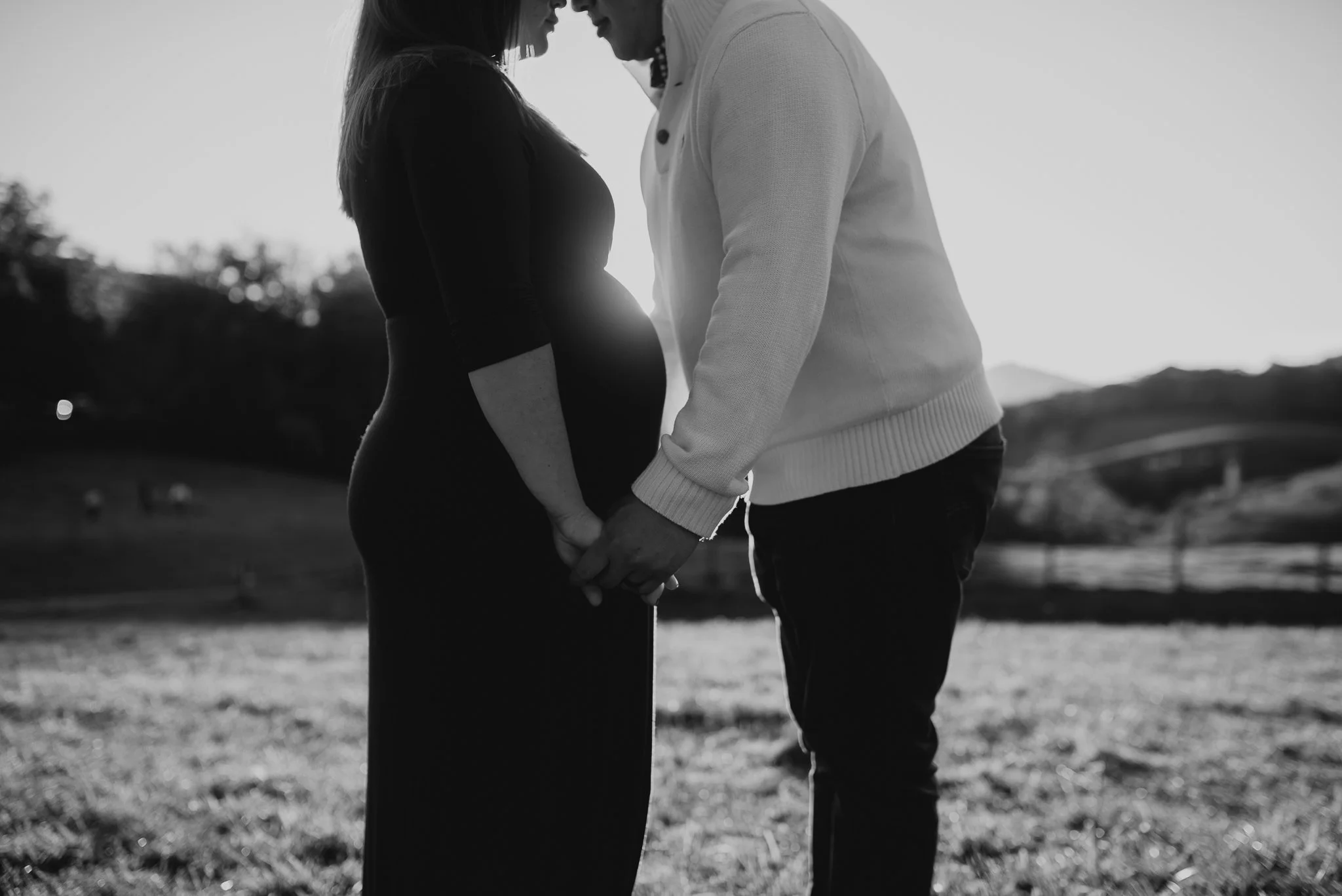 A black and white photo of a couple standing close, holding hands, with their foreheads touching, outdoors in a park or field during sunset.
