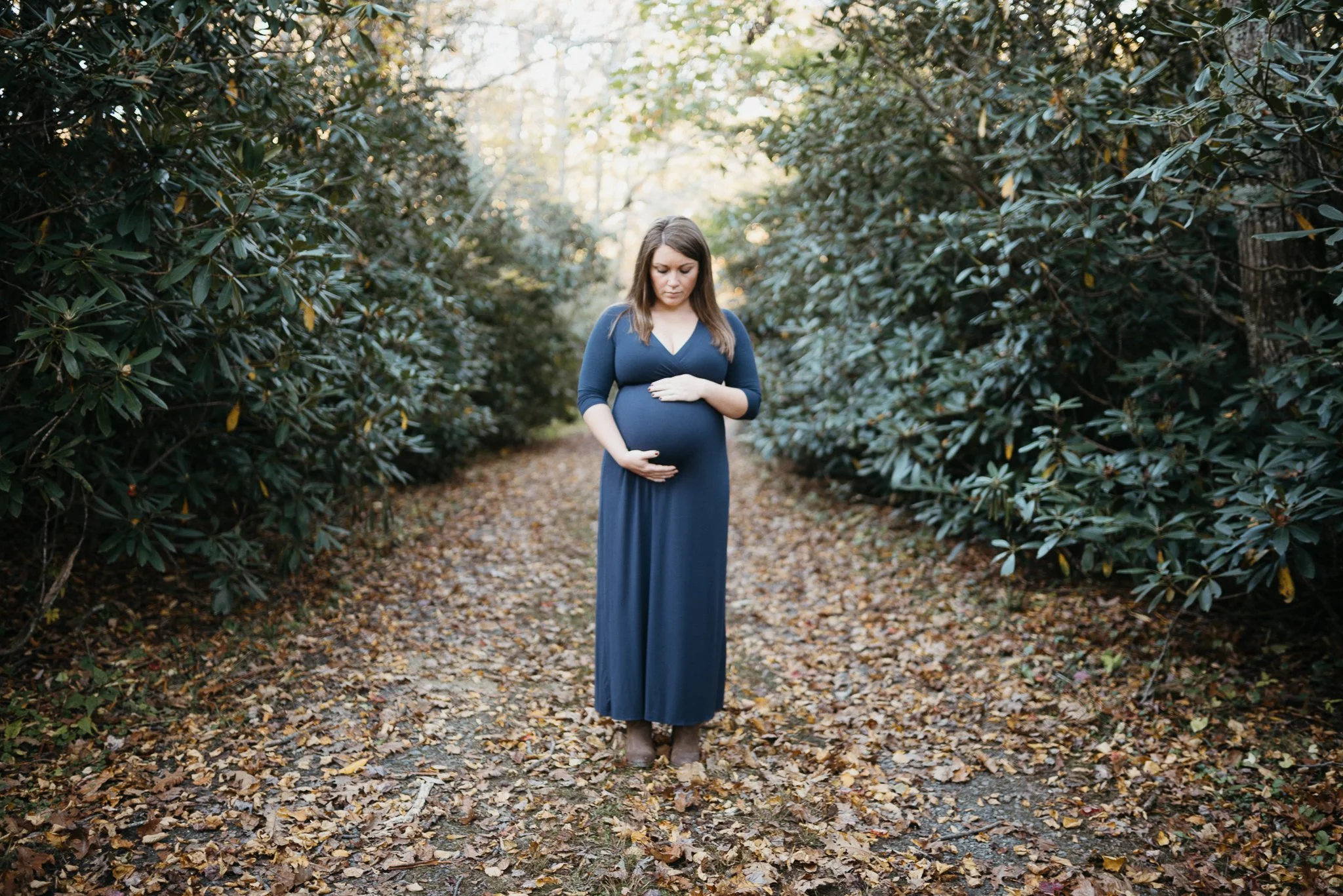 A pregnant woman in a long blue dress is standing on a forest path surrounded by green foliage, holding her belly with both hands, looking down with a thoughtful expression.