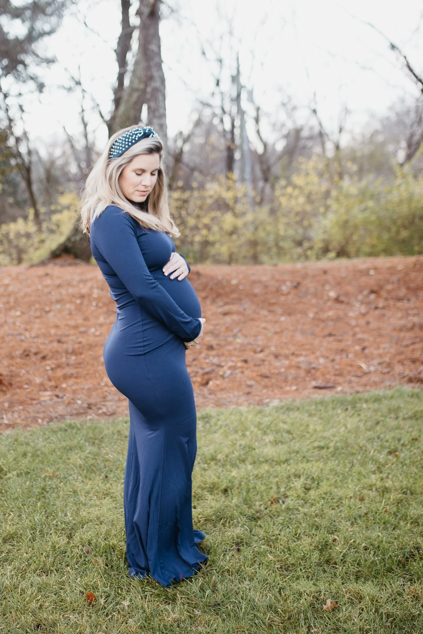 Pregnant woman in a blue dress with long sleeves standing outdoors in a park surrounded by trees with fall foliage, looking down at her baby bump with her hands resting on her stomach.