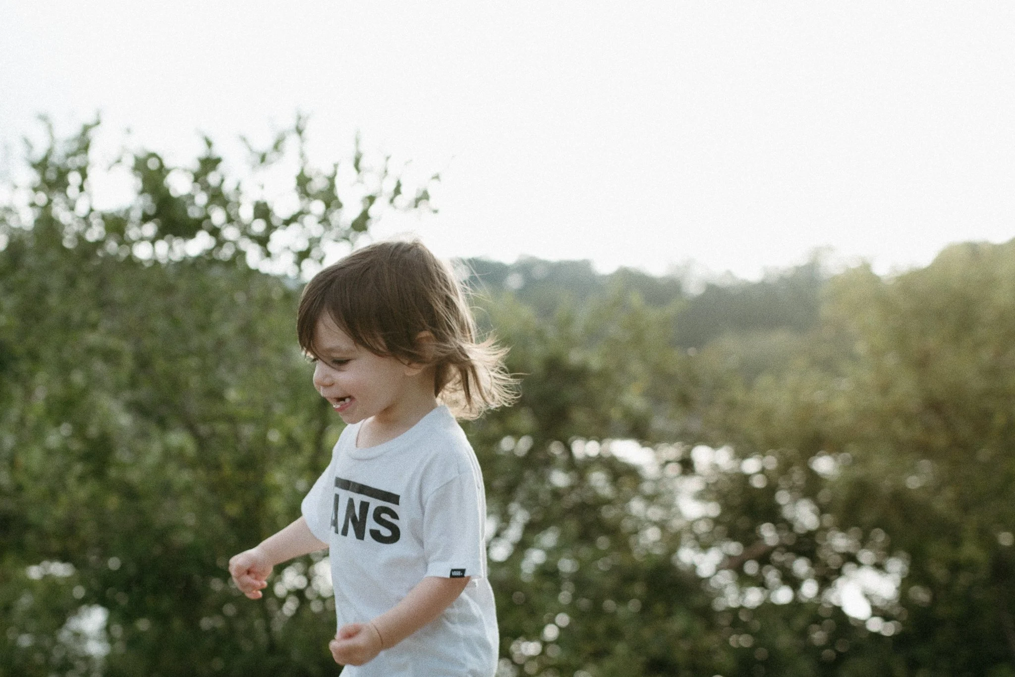 A young boy with light brown hair and a white t-shirt smiling and playing outdoors among green trees in a park or natural setting.