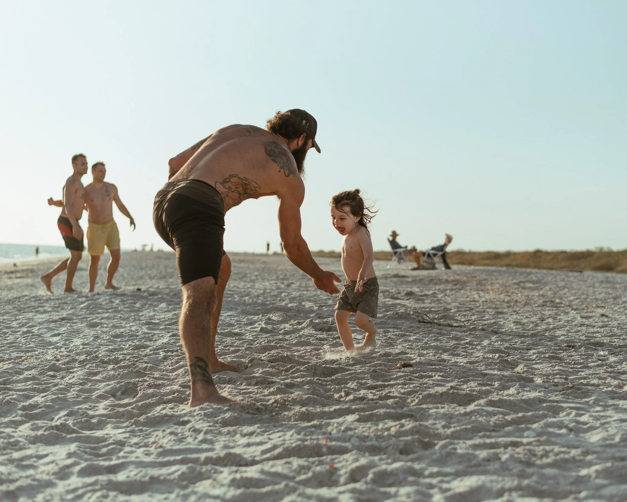 A man and a young girl playing on the sandy beach during sunset, with other beachgoers relaxing in the background.