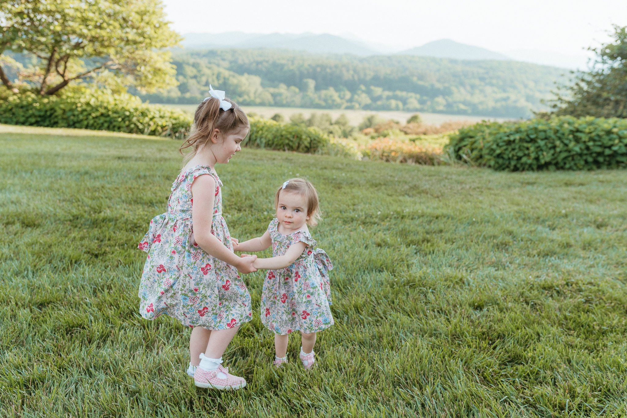 Two young girls in matching floral dresses holding hands and playing outdoors on a grassy field with trees and mountains in the background.