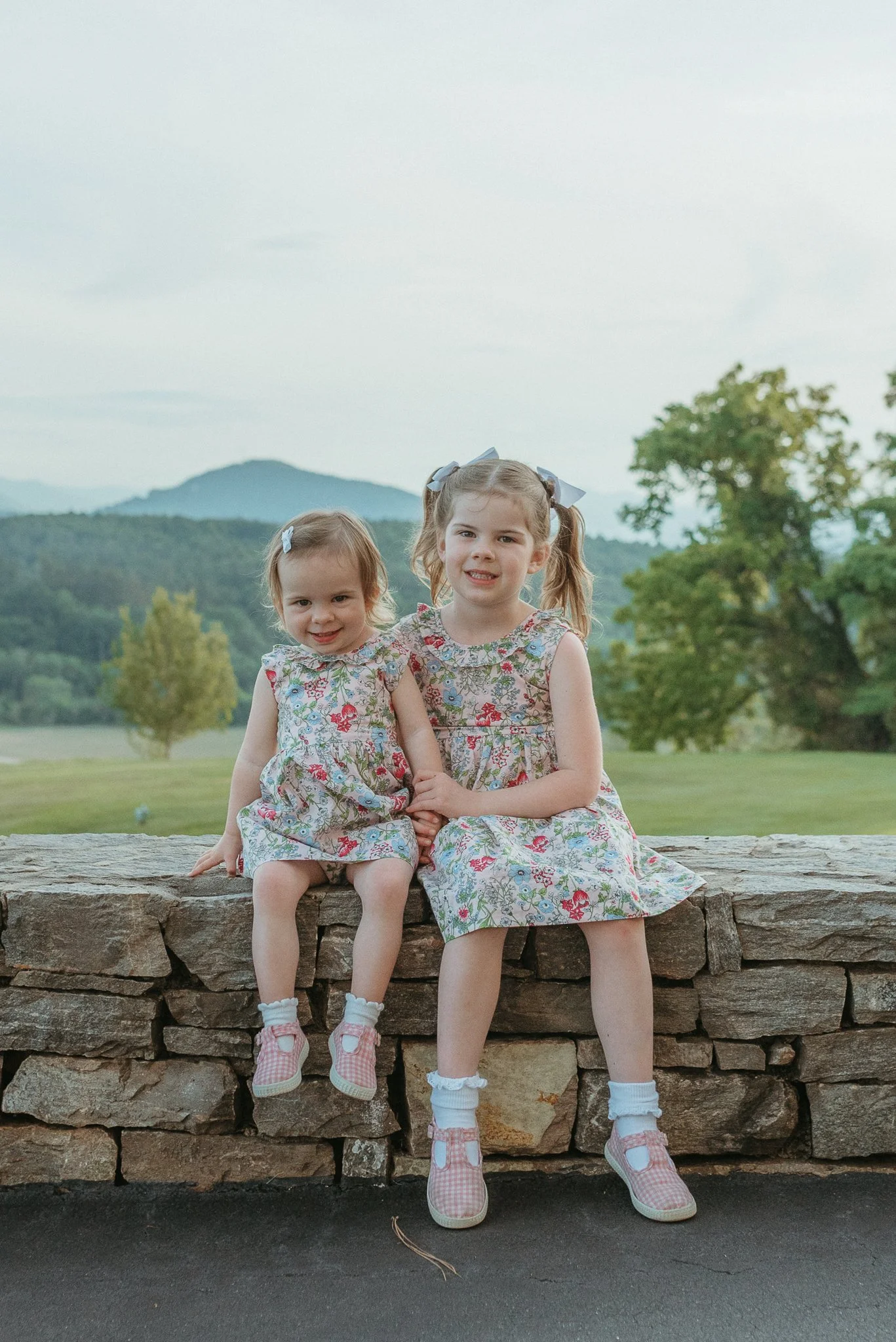 Two young girls sitting on a stone wall outdoors with trees and mountains in the background, wearing matching floral dresses and pink shoes, smiling at the camera.