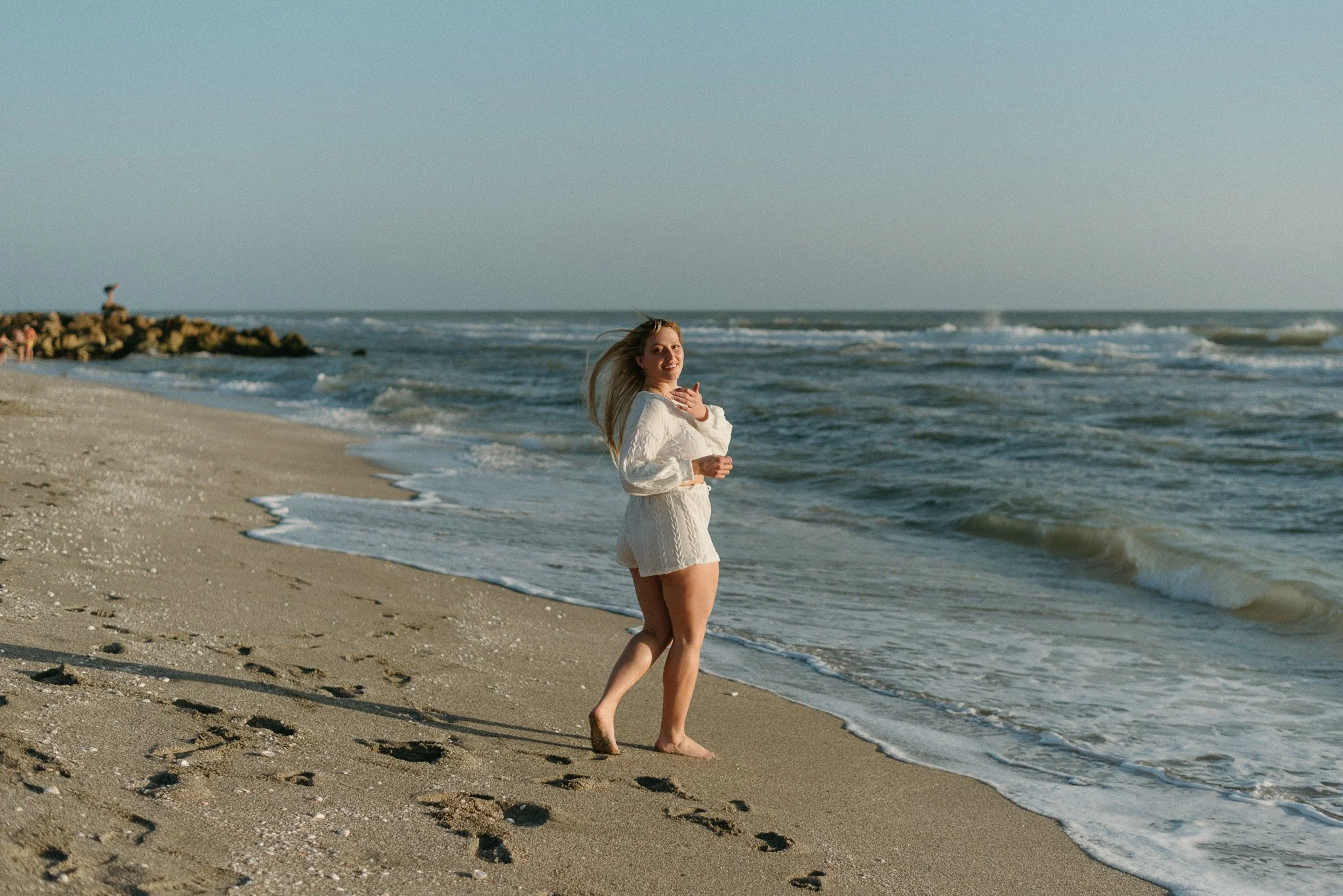 A woman in a white dress walking along the shoreline at the beach, with the ocean waves behind her and footprints on the sand.