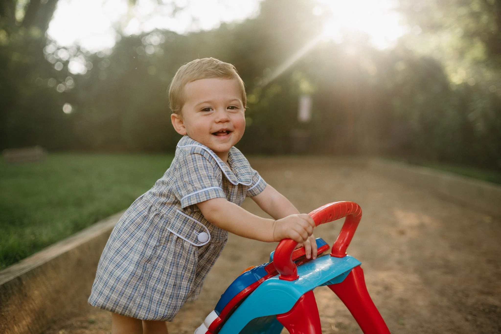 A young boy smiling while riding a tricycle outdoors in a park during late afternoon sunlight.